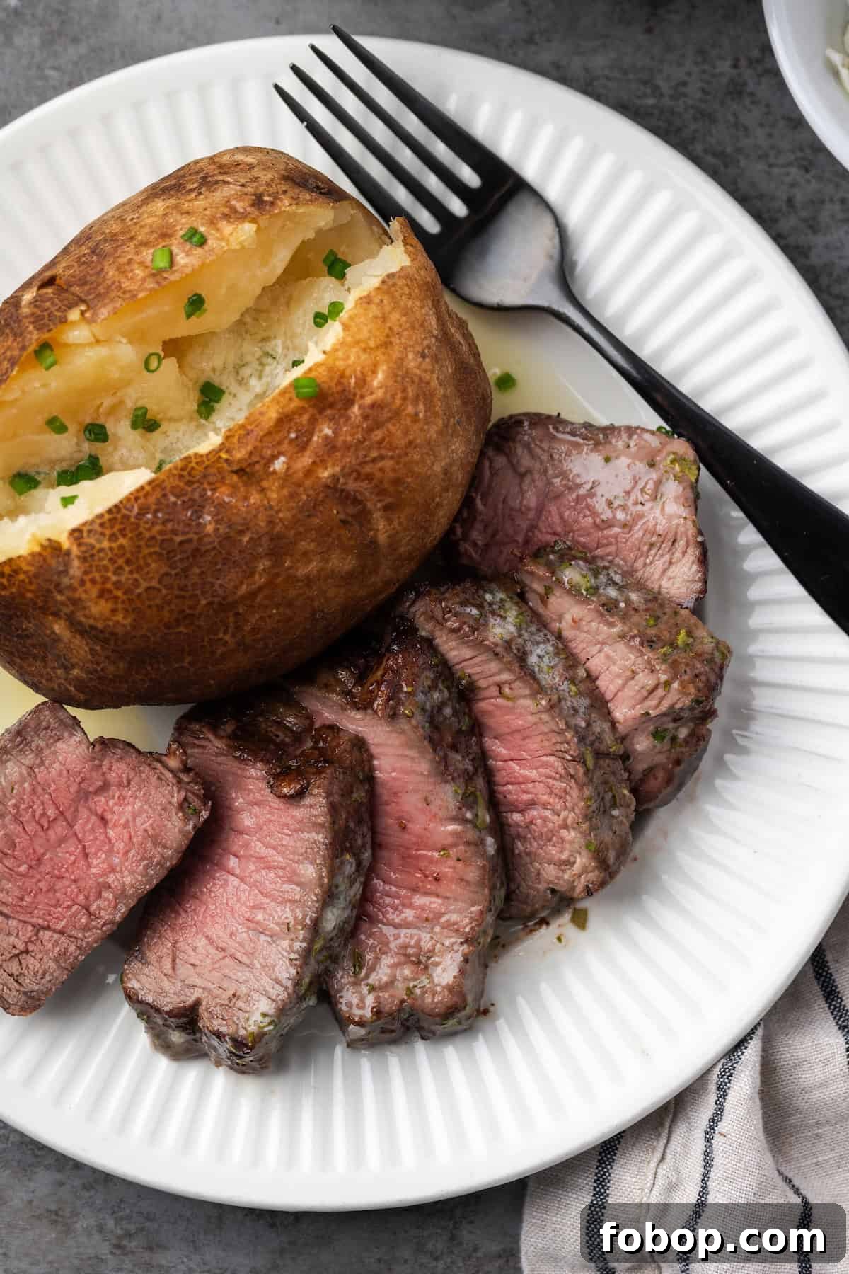 Overhead view of air fryer filet mignon cut into slices next to a baked potato and a fork on a plate.