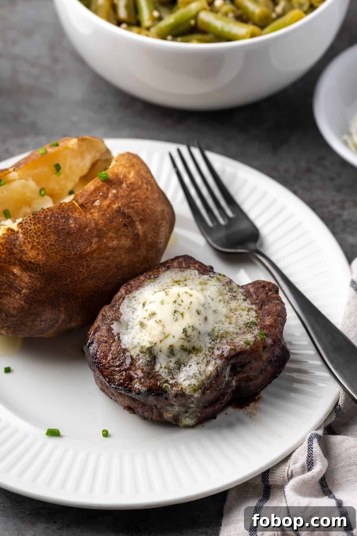 Air fryer filet mignon topped with garlic butter next to a baked potato on a plate, with a fork in the background.