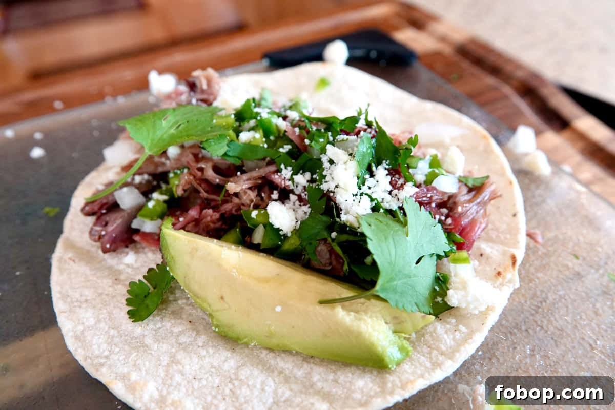 Smoked Pulled Pork Tacos with Tangy Pineapple Slaw 2 White corn tortilla topped with shredded smoked pork and chopped white onion and jalapeños, white crumbled cheese and cilantro leaves, and a slice of avocado on glass cutting board with wood cutting board underneath.