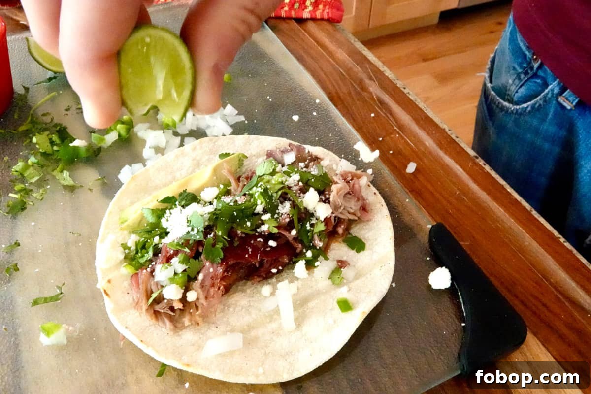 Smoked Pulled Pork Tacos with Tangy Pineapple Slaw 7 lime slice squeeze by hand, on top of smoked pull pork on a white corn tortilla garnished with crumbled white cheese, chopped up white onion and jalapeño, chopped cilantro and a slice of avocado on top of a glass board.