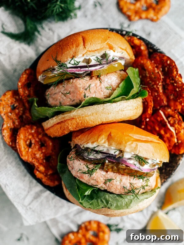 Two salmon burgers in a serving dish stacked with lettuce, pickles, onions, and mayo, with side of pretzels.