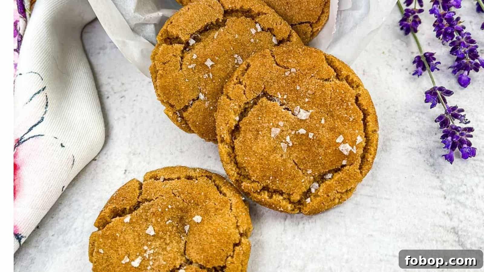 25 Office Cookies Everyone Brings and Raves About 16 Three brown butter snickerdoodle cookies arranged on a white surface, sprinkled with sea salt flakes, with a white cloth and purple flowers on the side.