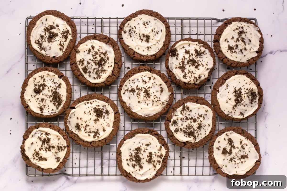 25 Office Cookies Everyone Brings and Raves About 4 A cooling rack with twelve Crumbl Copycat Frosted Oreo Cookies topped with white frosting and chocolate crumbs, arranged on a white marble surface.