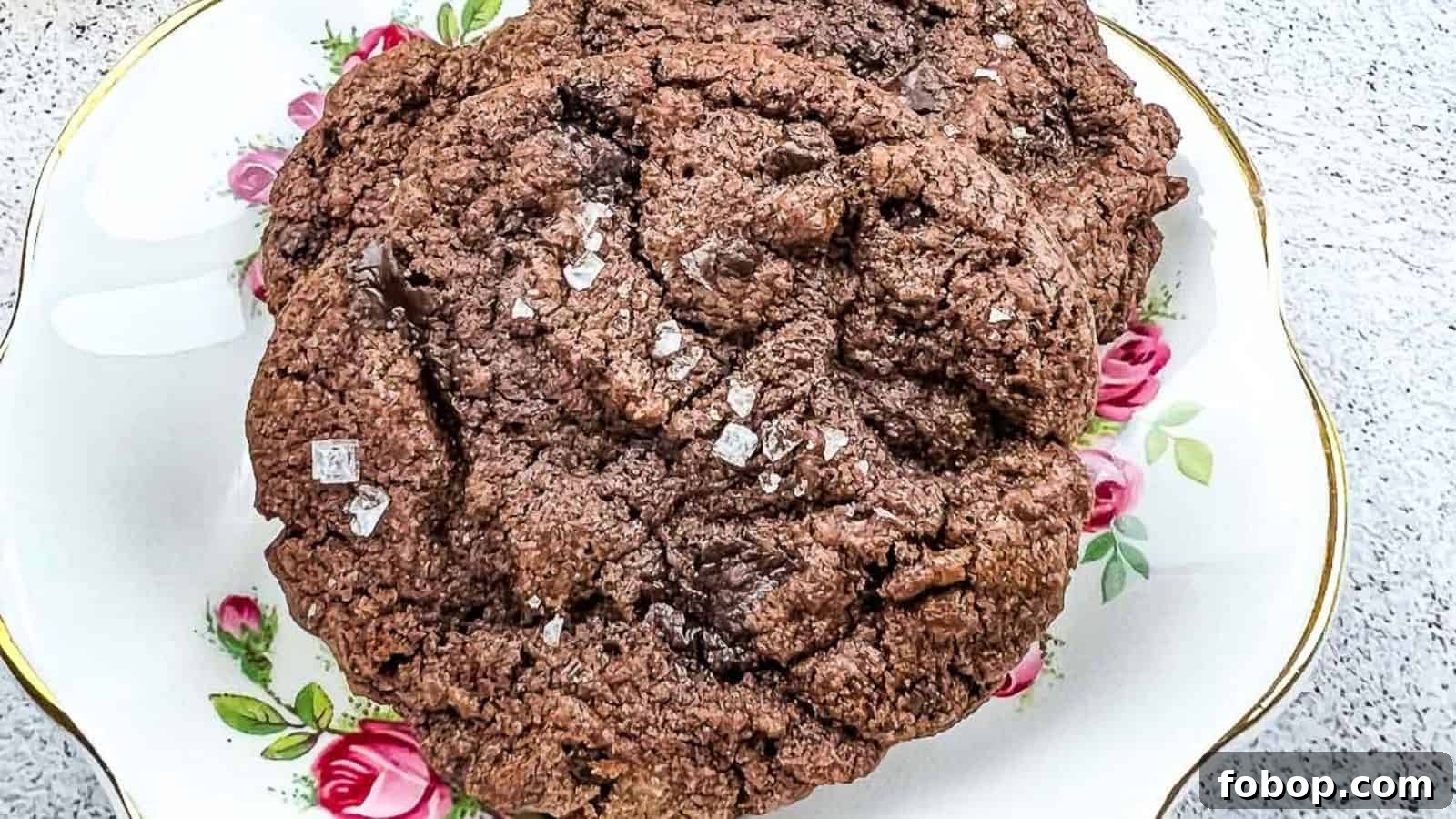 25 Office Cookies Everyone Brings and Raves About 5 A close-up of a large chocolate cookie sprinkled with coarse salt, placed on a decorative plate with pink rose patterns.