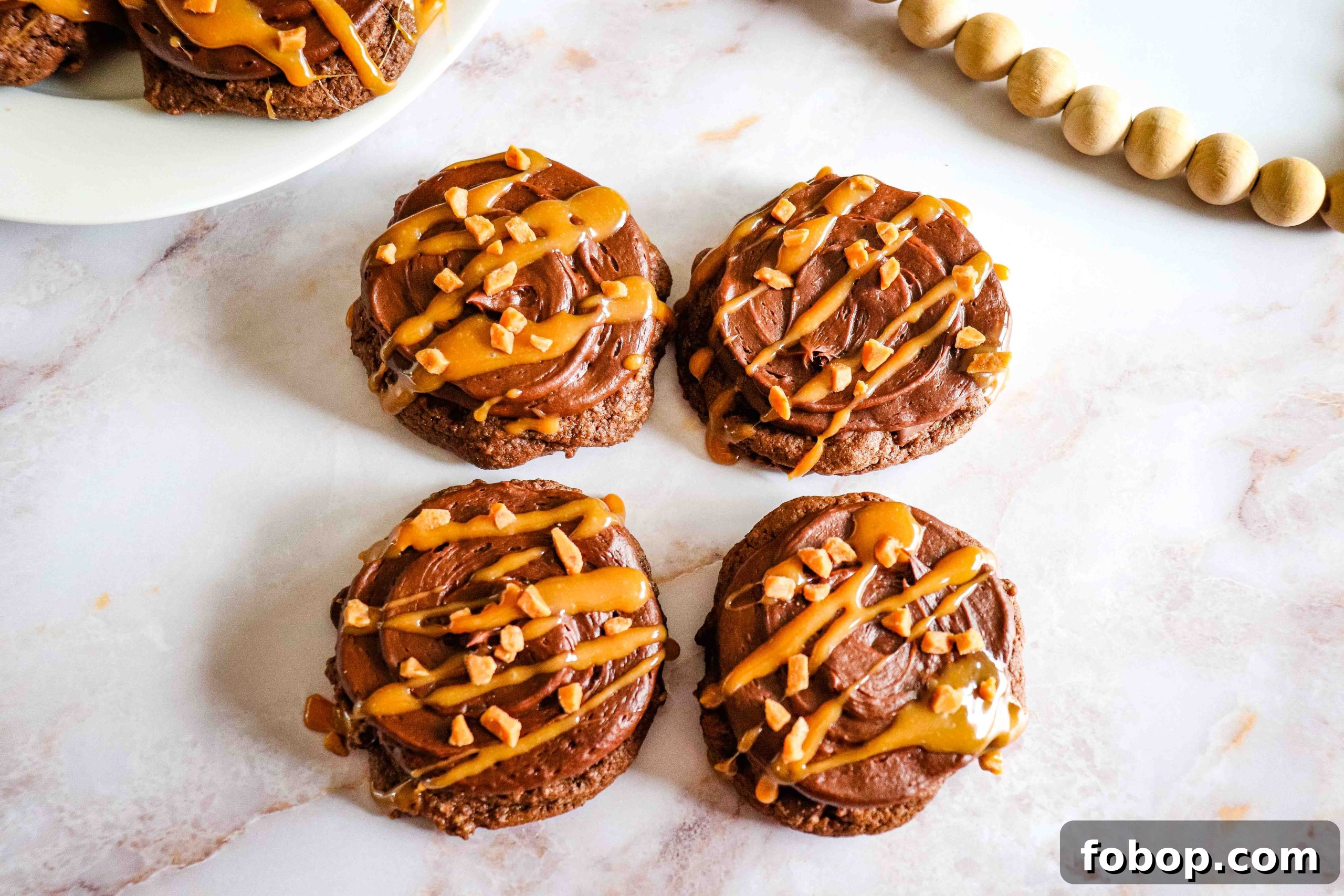 25 Office Cookies Everyone Brings and Raves About 9 Chocolate Caramel Cookies with frosting, caramel drizzle, and chopped nuts on a white marble surface.