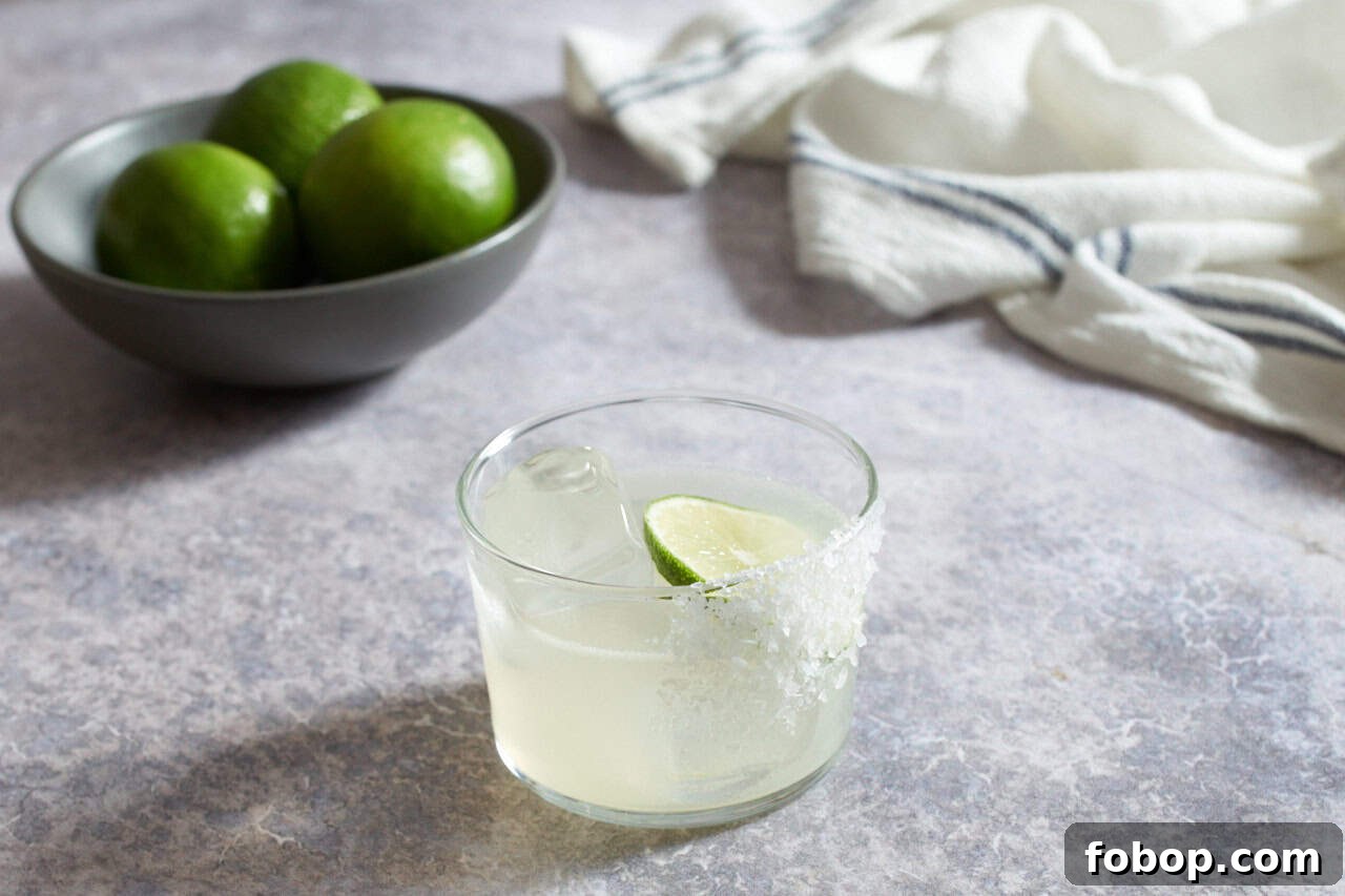 Smoky Mezcal Margarita Recipe: How to Make the Classic Cocktail 5 A mezcal margarita with salt, a bowl of limes and a blue and white striped towel are in the background.