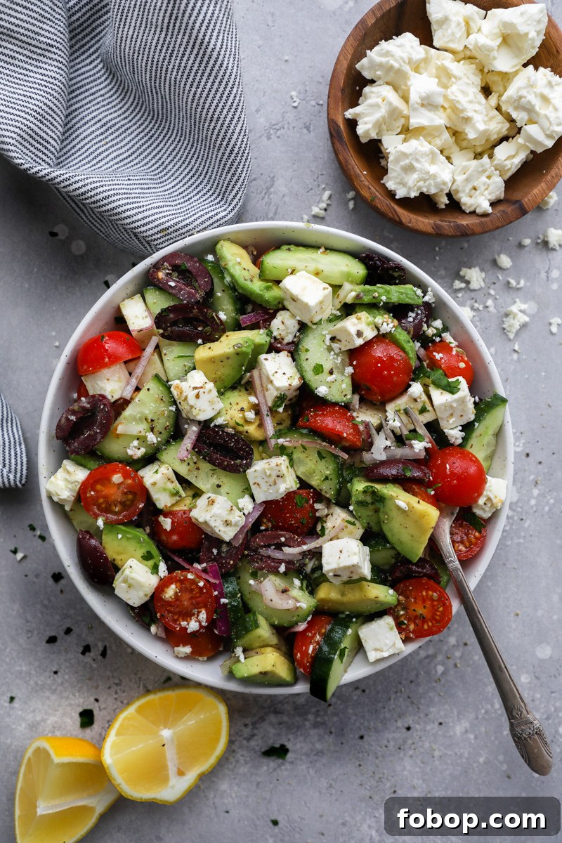 A no lettuce salad with avocado on a white plate surrounded by lemon wedges and a small bowl of crumbled feta cheese