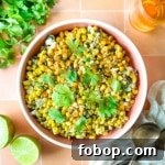 Overhead shot of an elote bowl surrounded by halved limes, cilantro, a cup of beer, and a white linen.