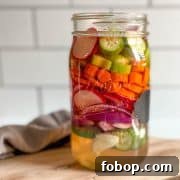 A glass jar of mixed pickles sits on a wooden cutting board with a gray linen.