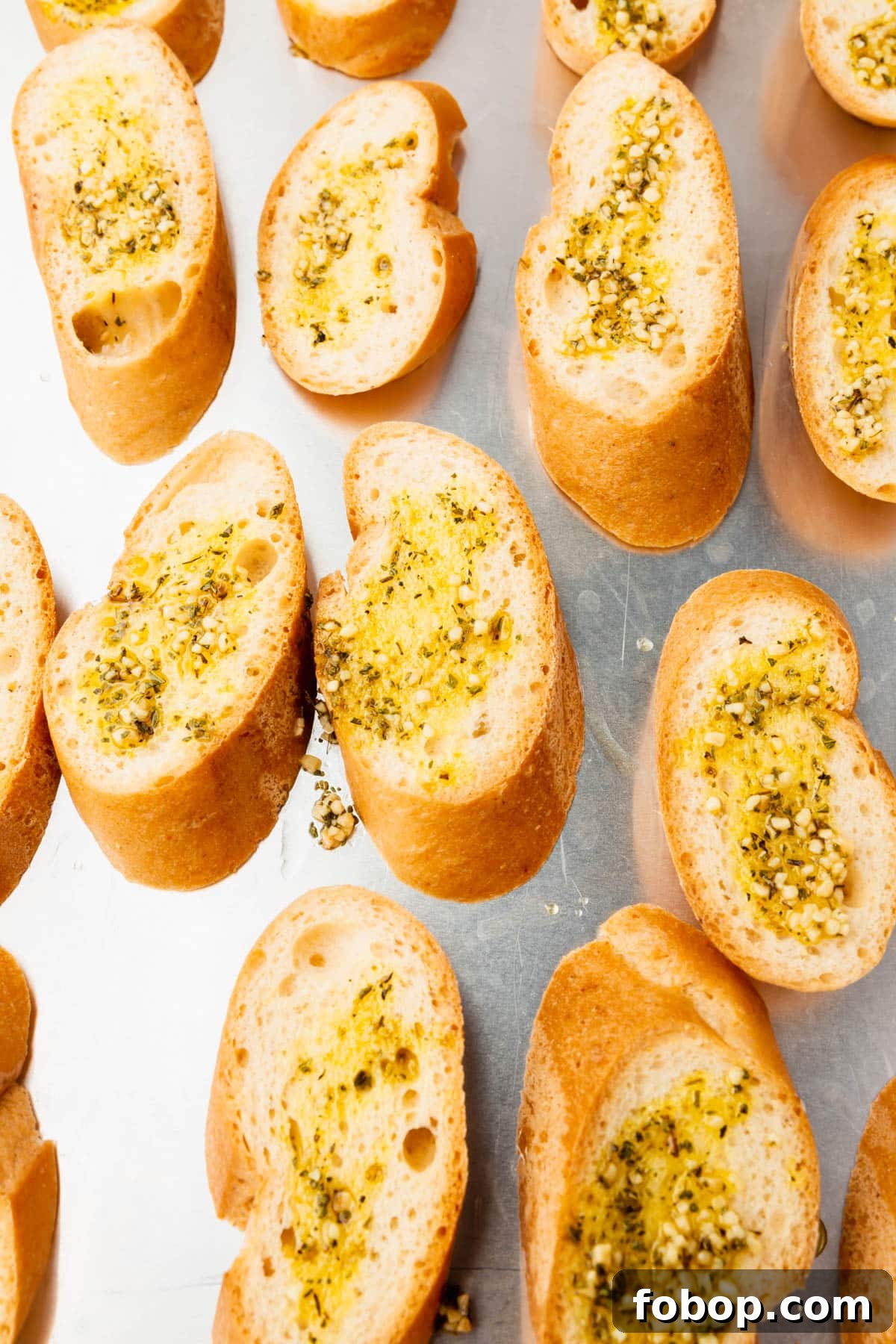 A close up of gluten free bread on a baking sheet topped with olive oil, minced garlic and dried herbs before toasting in the oven.