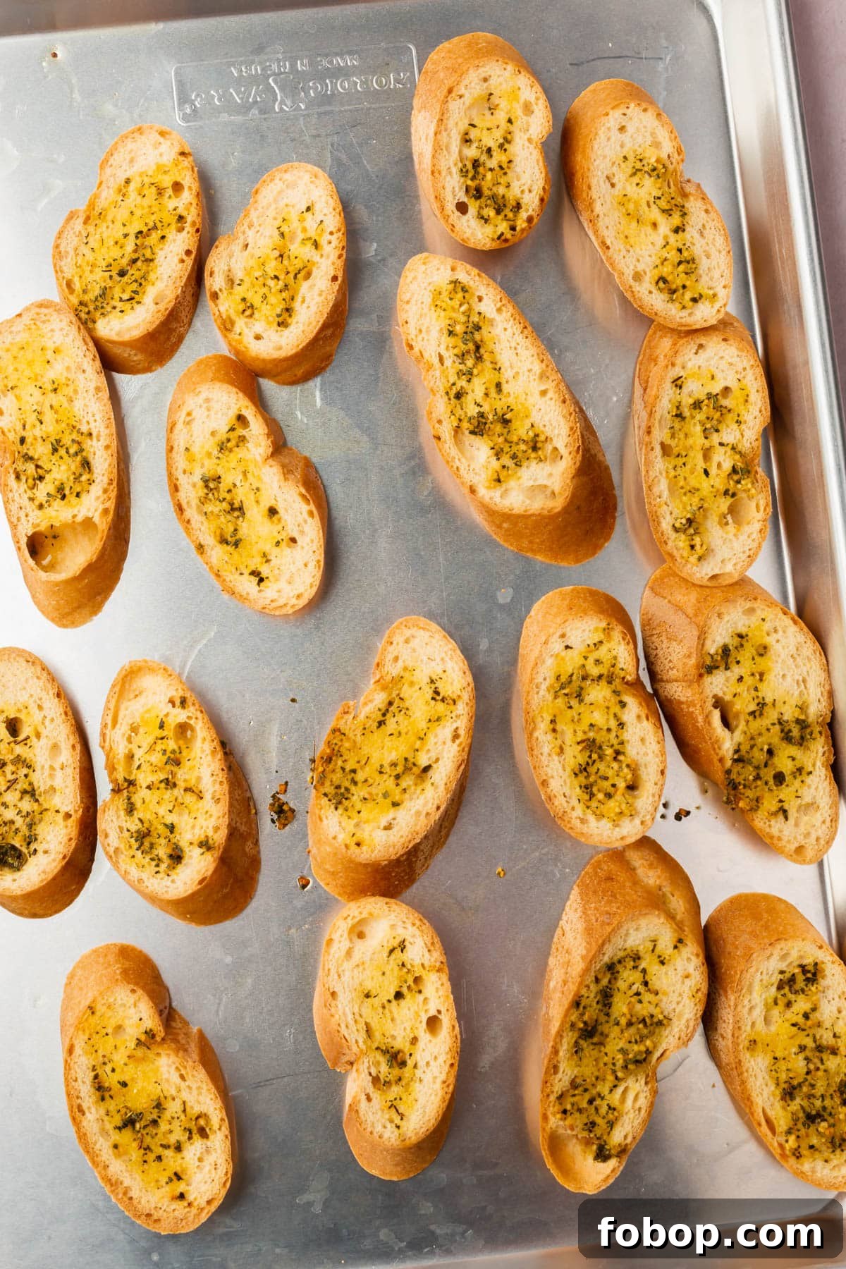 A sheet pan with slices of gluten-free bread cut on the bias and topped with olive oil, minced garlic and dried herbs.