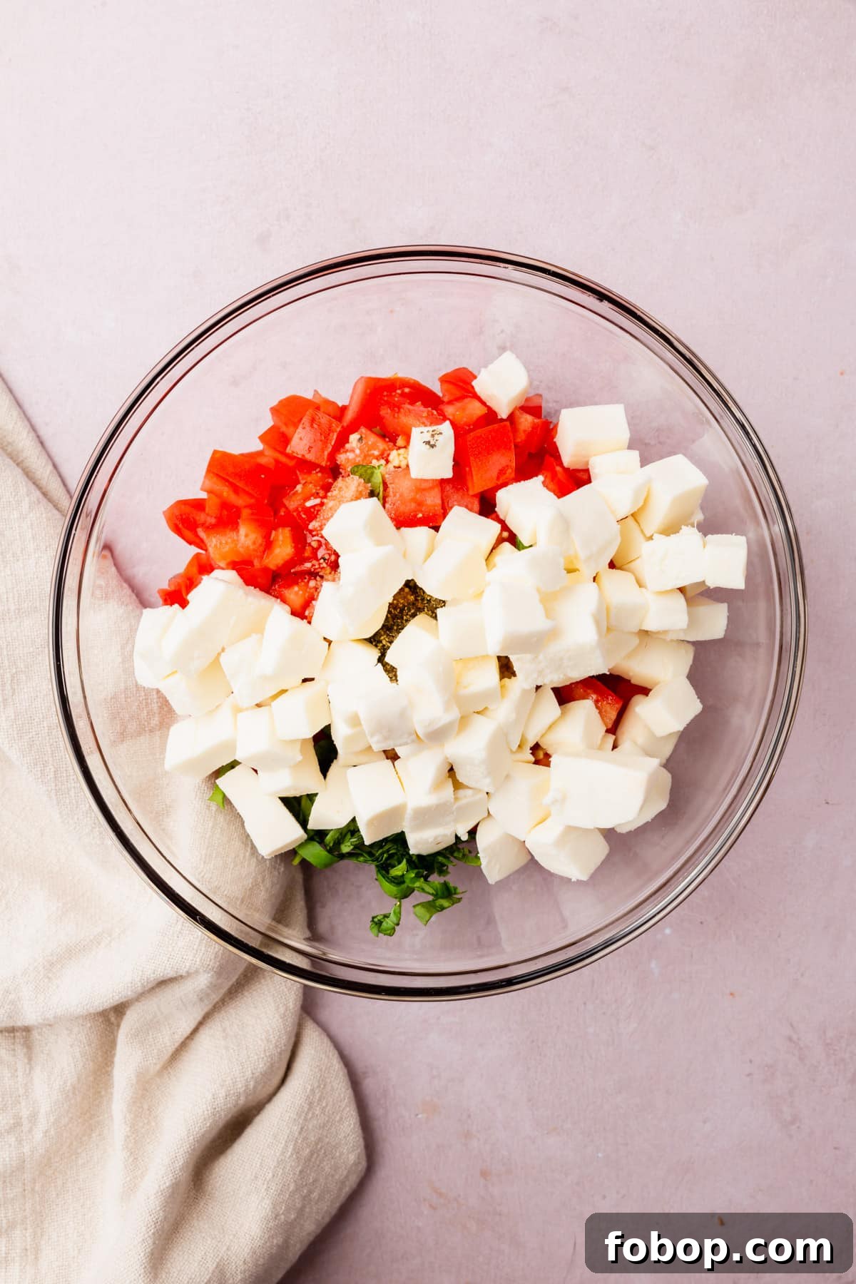 A glass mixing bowl with diced tomatoes, sliced basil, diced fresh mozzarella and minced garlic before mixing together.