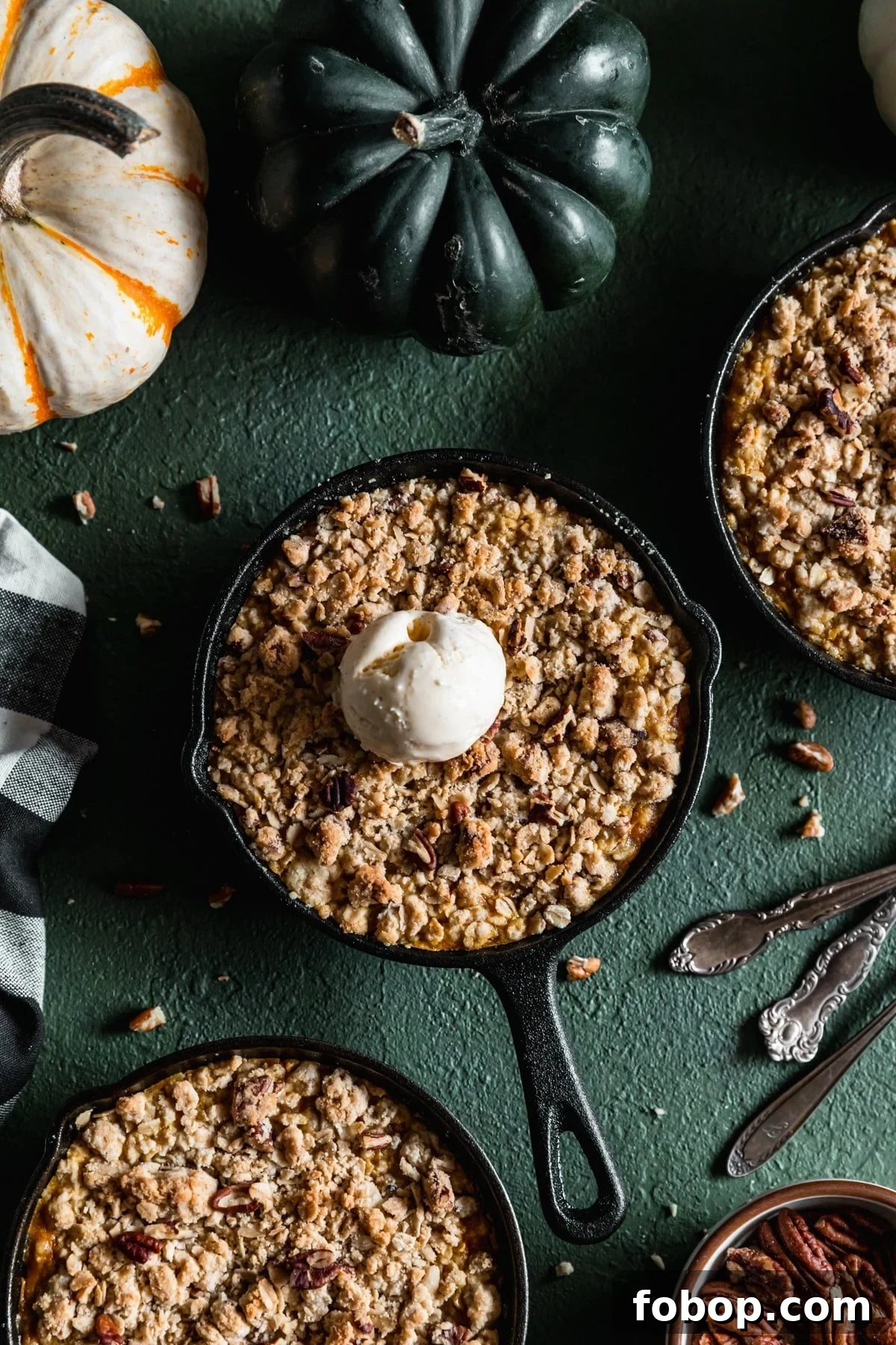 Maple Oat Streusel Pumpkin Crisp Recipe 2 An overhead image of a mini cast iron skillet with pumpkin crisp and a scoop of vanilla ice cream on a dark green table next to two more skillets, a black and white checkered linen, and vintage forks.