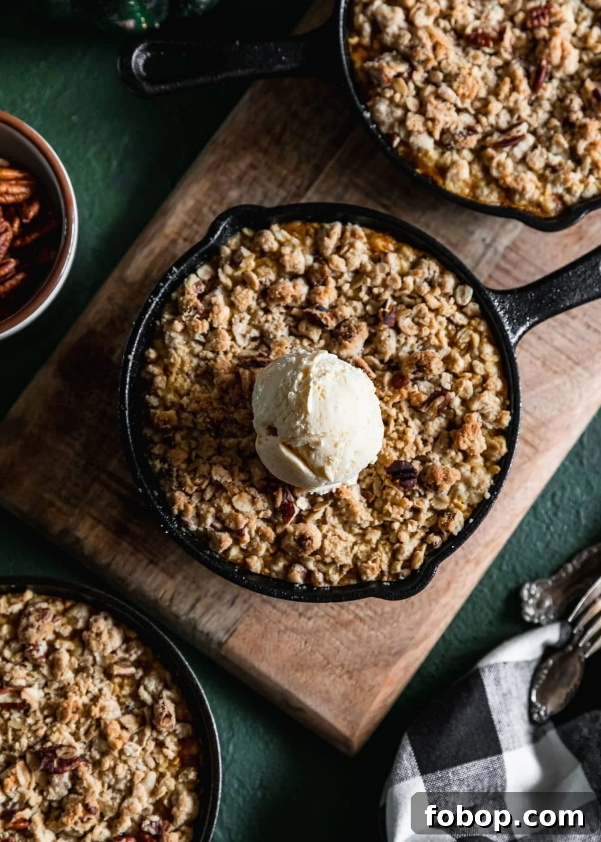 Maple Oat Streusel Pumpkin Crisp Recipe 8 A closeup of a mini cast iron skillet with pumpkin crisp and a scoop of vanilla ice cream on a wood board next to another crisp. The board is on a green table next to a white bowl of pecans, a black and white linen, and vintage forks.