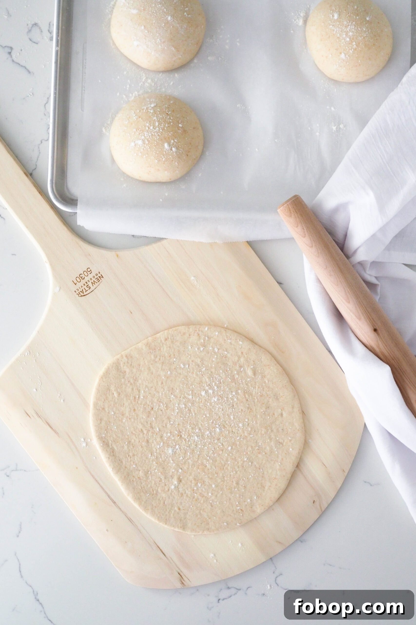 A rolled out pizza crust on a pizza peel, with three small dough balls and a rolling pin beside it.