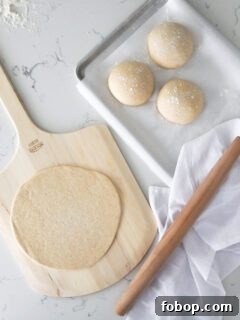 A rolled out pizza crust on a pizza peel, with three small dough balls and a rolling pin beside it.