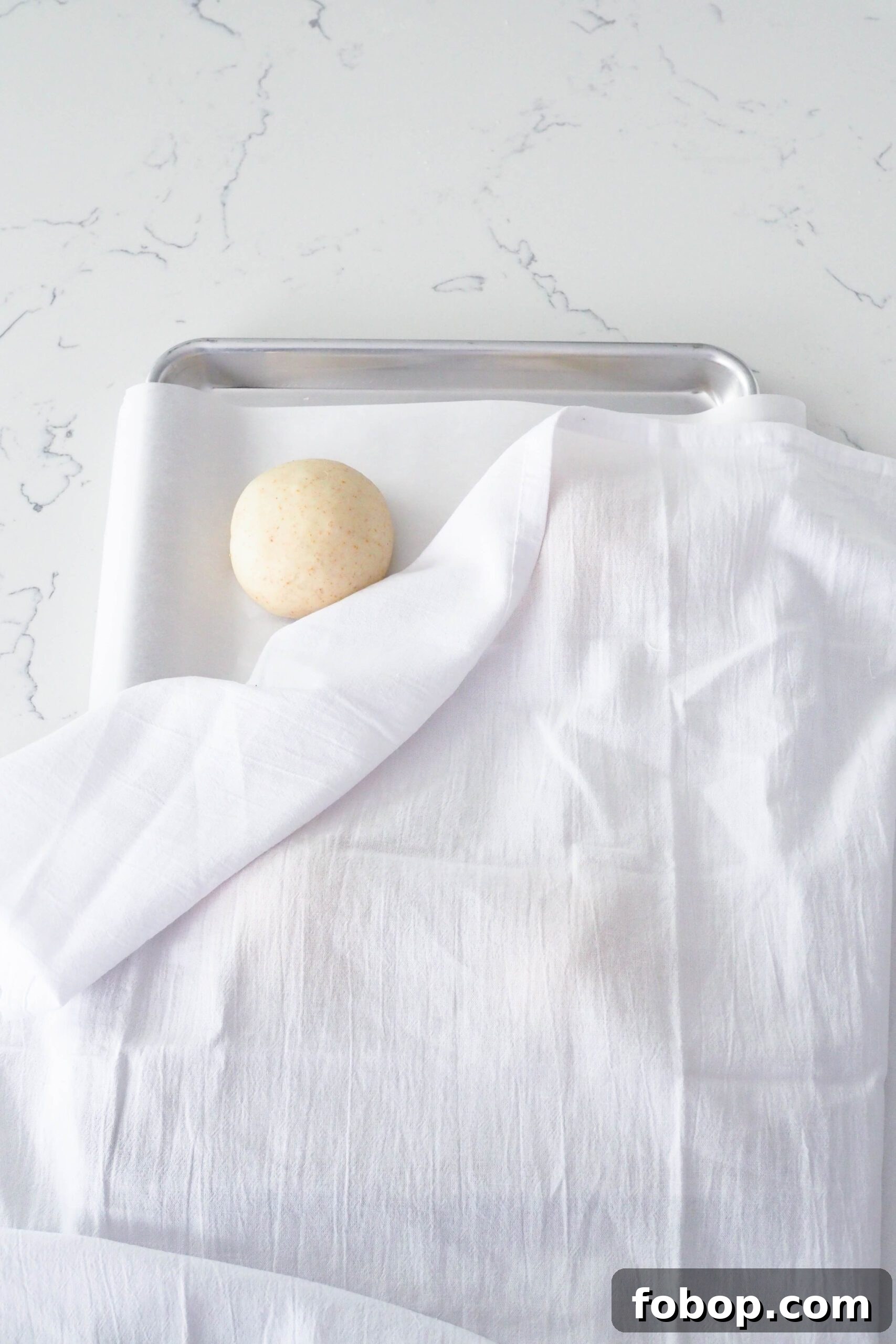 A dough ball peeks out behind a white kitchen towel on a baking sheet.
