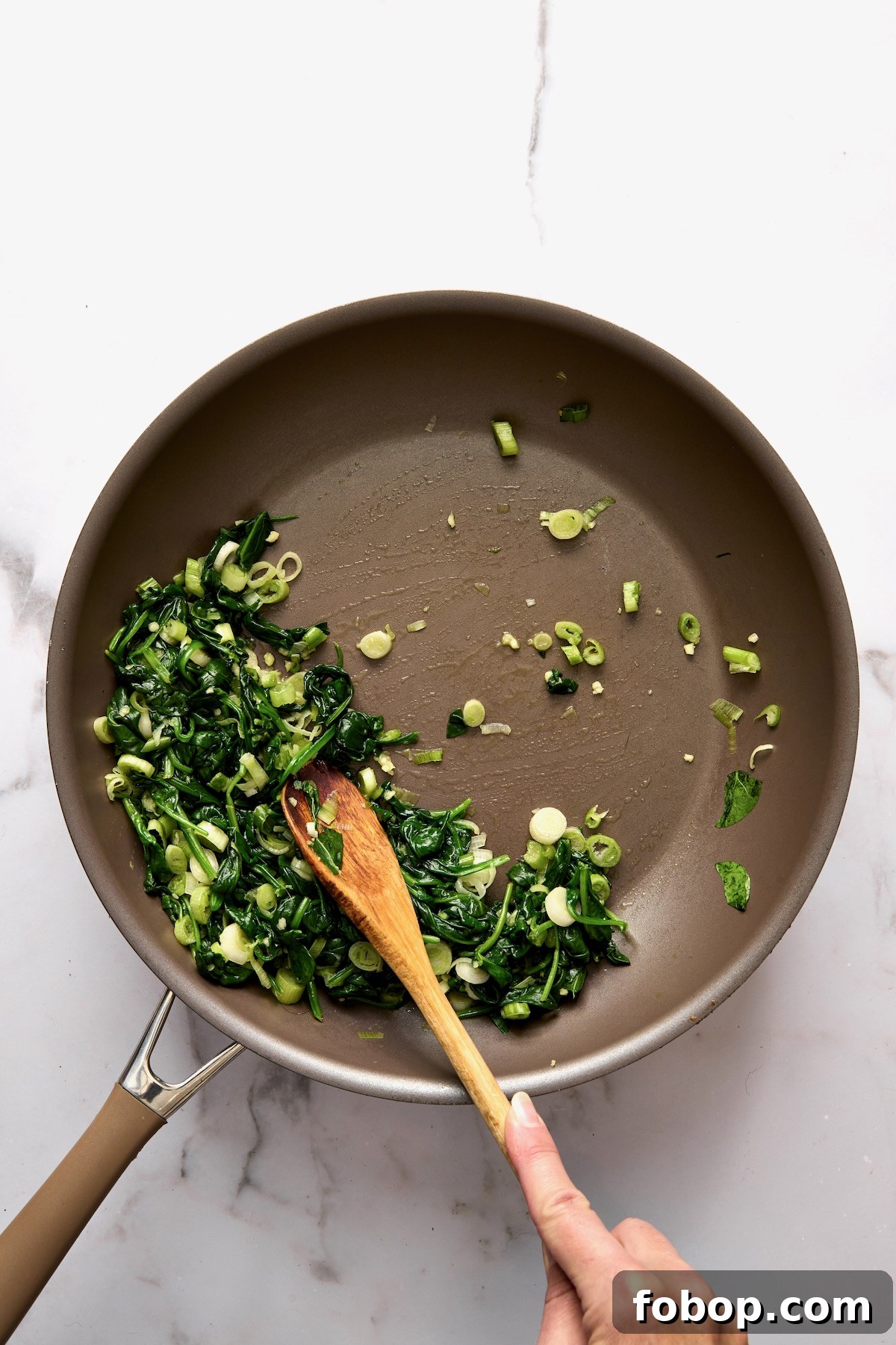 Cooked spinach and scallions in skillet being stirred with wooden spoon.