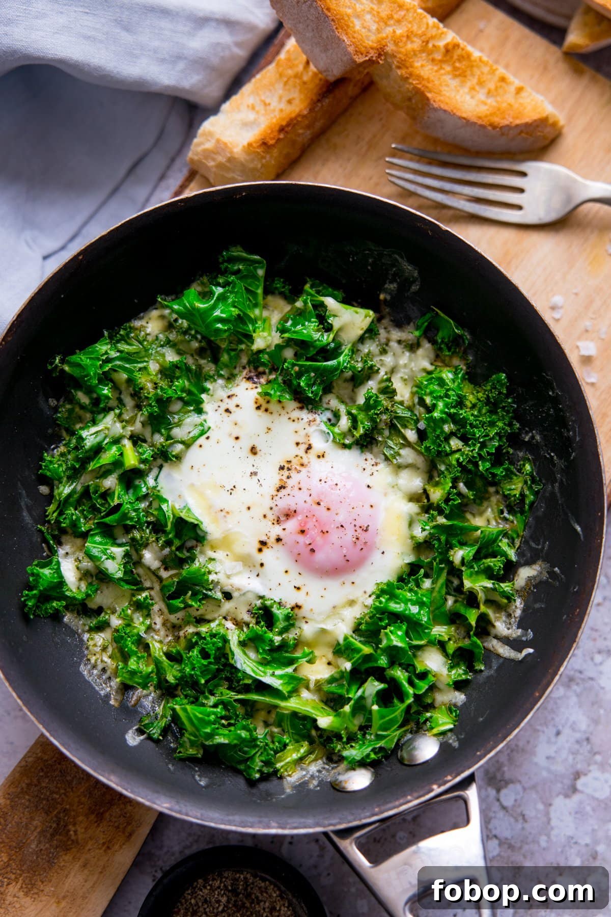 Creamy Scrambled Eggs with Sautéed Kale Breakfast Bowl 3 Fried egg in a pan with kale and cheese. Pan is on a wooden board with toast soldiers at the top of the shot.