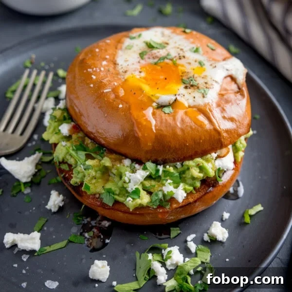 Creamy Scrambled Eggs with Sautéed Kale Breakfast Bowl 9 A bagel with a fried egg, smashed avocado, crumbled cheese, and chopped herbs sits on a dark plate with a fork beside it.
