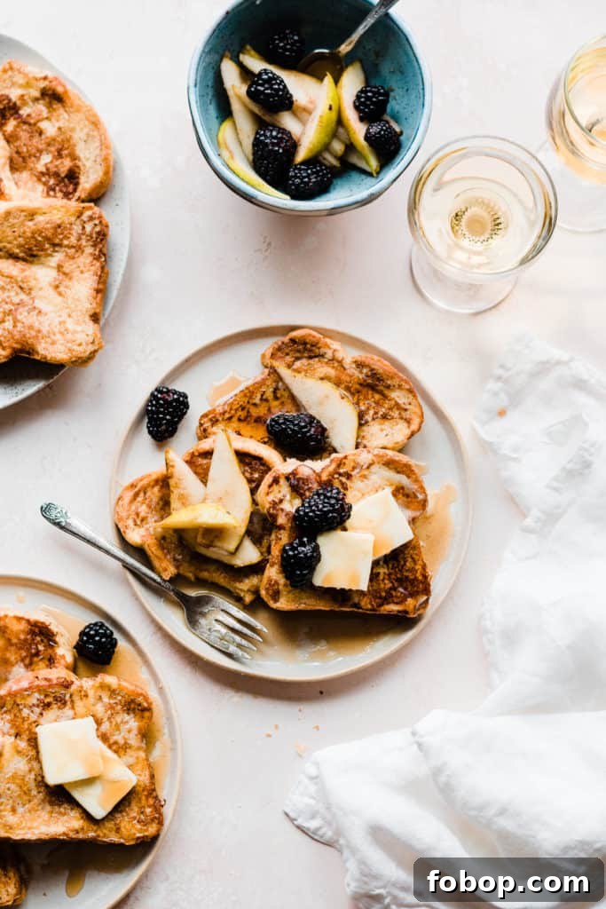 Two plates of golden french toast topped with butter, maple syrup, and blackberries and pear slices, on a light pink surface with two glasses of apple juice and a white linen