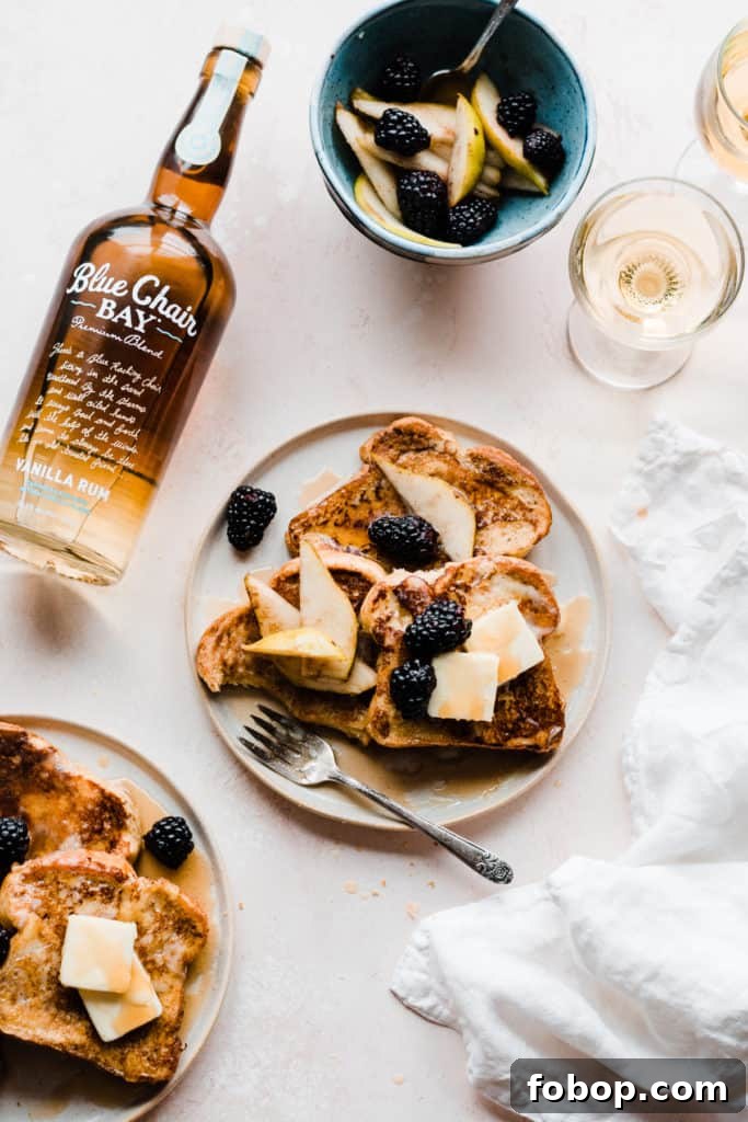 Two plates of golden french toast topped with butter, maple syrup, and blackberries and pear slices, on a light pink surface with two glasses of apple juice and a white linen. A bottle of Blue Chair Bay Vanilla Rum is laying next to the plates