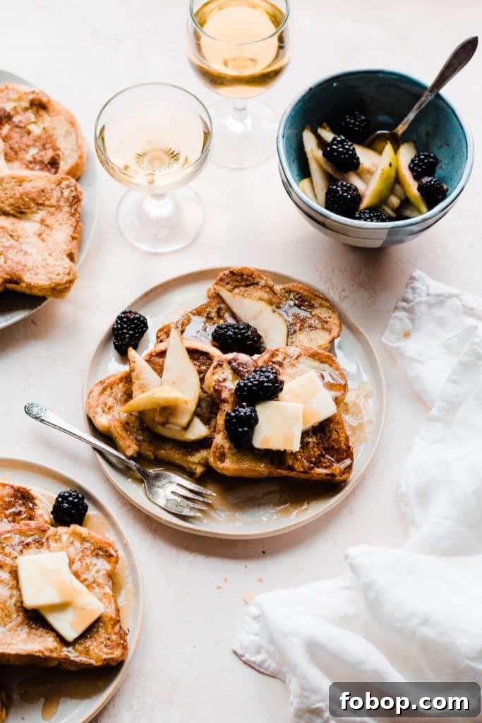 Two plates of golden french toast topped with butter, maple syrup, and blackberries and pear slices, on a light pink surface with two glasses of apple juice and a white linen. A bite has been taken from the french toast