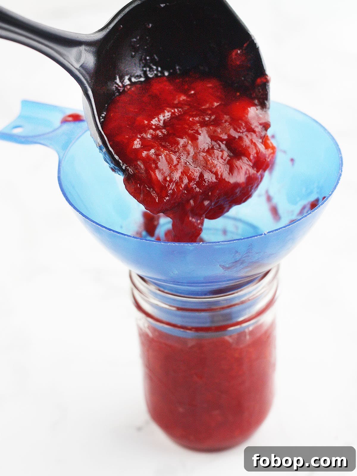 ladling strawberry jam into a mason jar with a blue wide mouth funnel