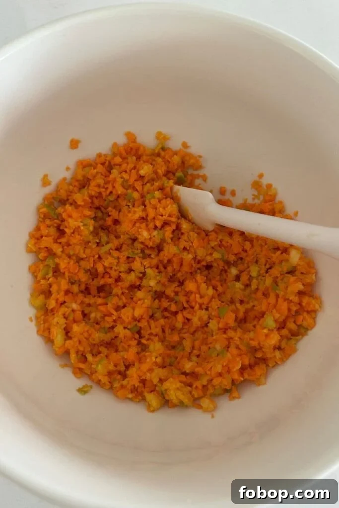 A bowl of grated carrot and apple being stirred together with a white jar spatula.