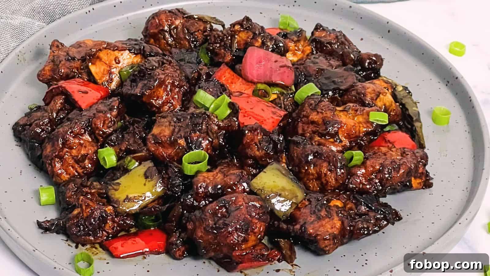 A plate of Chilli Chicken with chunks of red peppers, drizzled in a dark sauce, garnished with green onions.