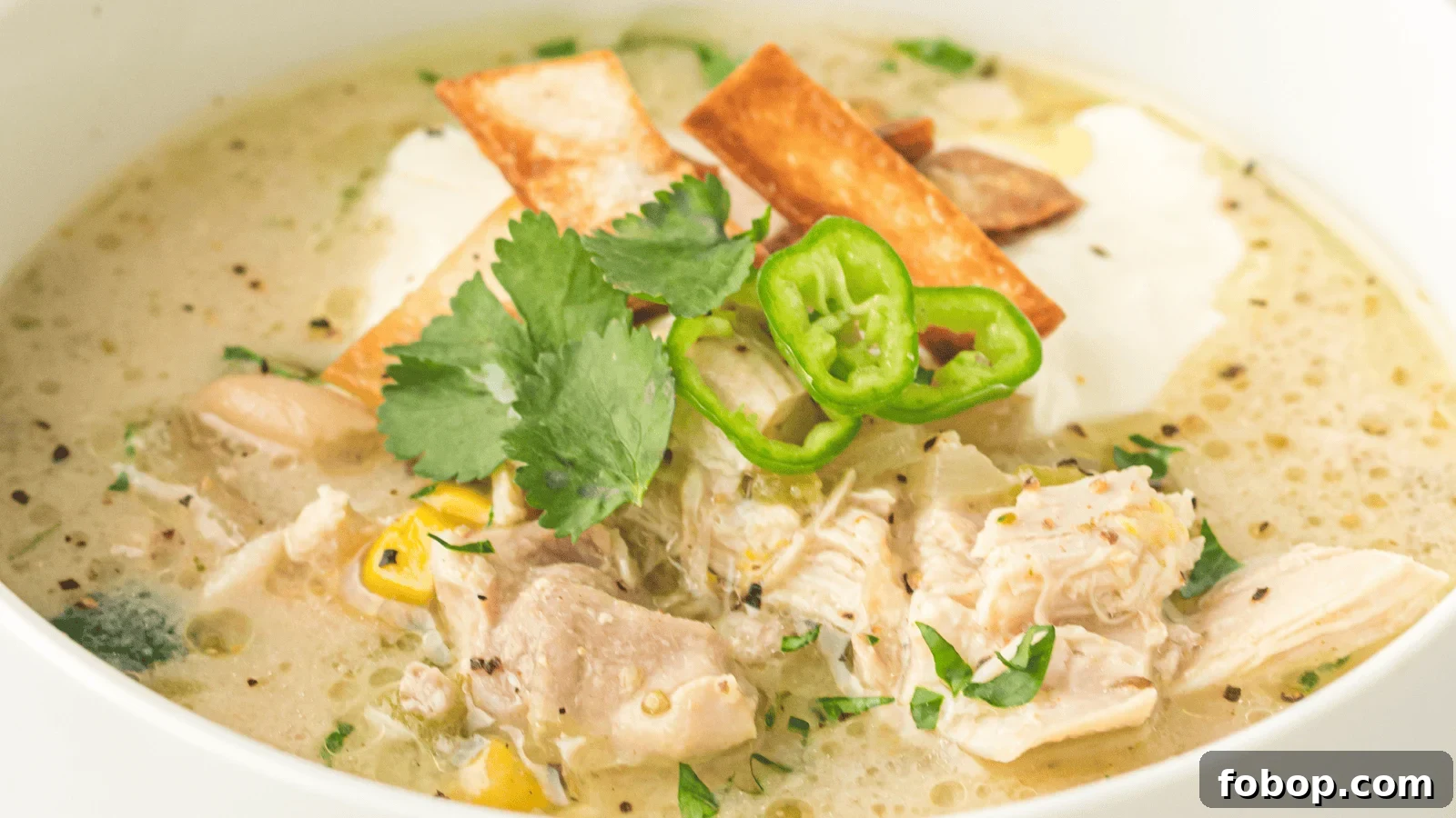 Bowl of creamy chicken soup topped with tortilla strips, sliced green jalapeños, cilantro, corn, and a dollop of sour cream.