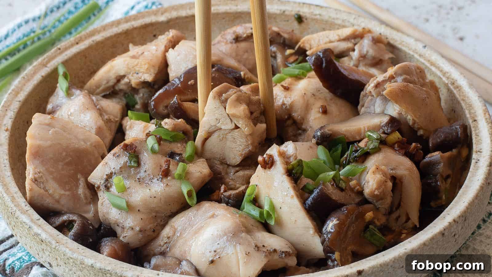 A bowl of steamed chicken pieces with mushrooms, garnished with chopped green onions, with chopsticks picking up a piece.
