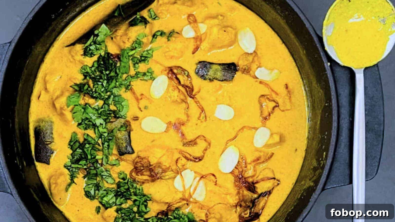 A bowl of Chicken Korma garnished with herbs and sliced almonds, served with a spoon on the side.