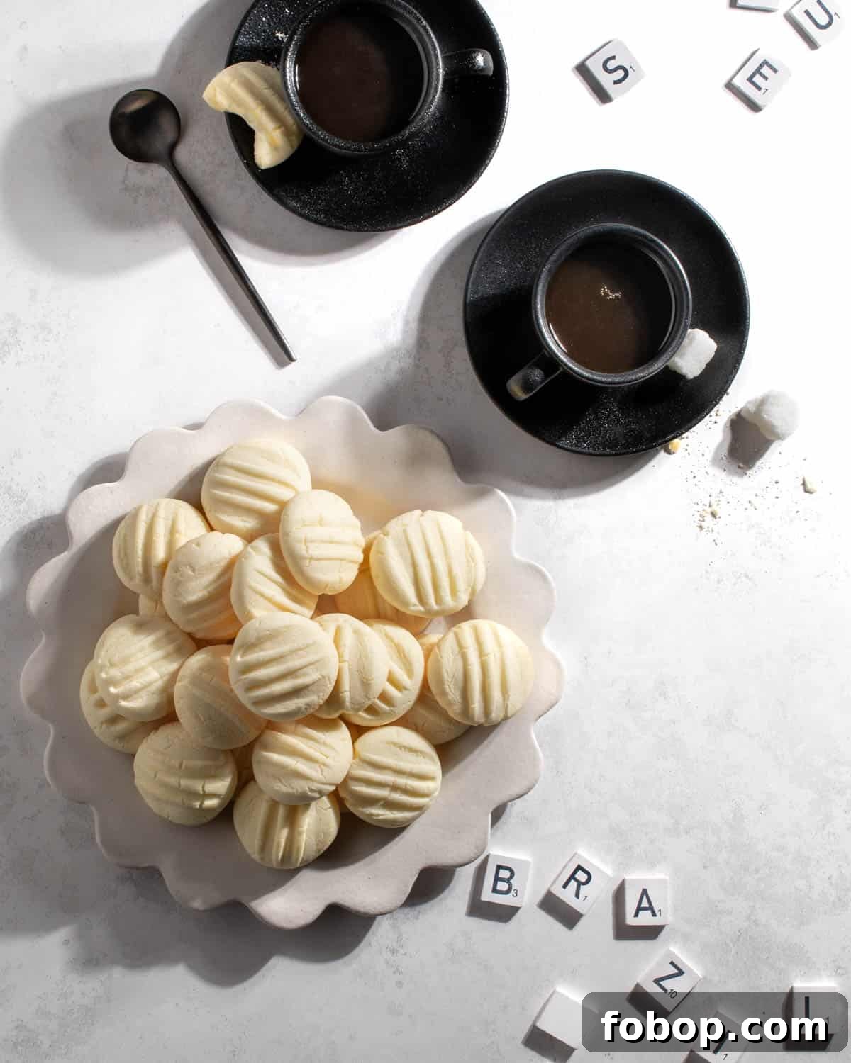 Sequilhos cookies on a white plate with two cups of coffee and some scrabble pieces. 