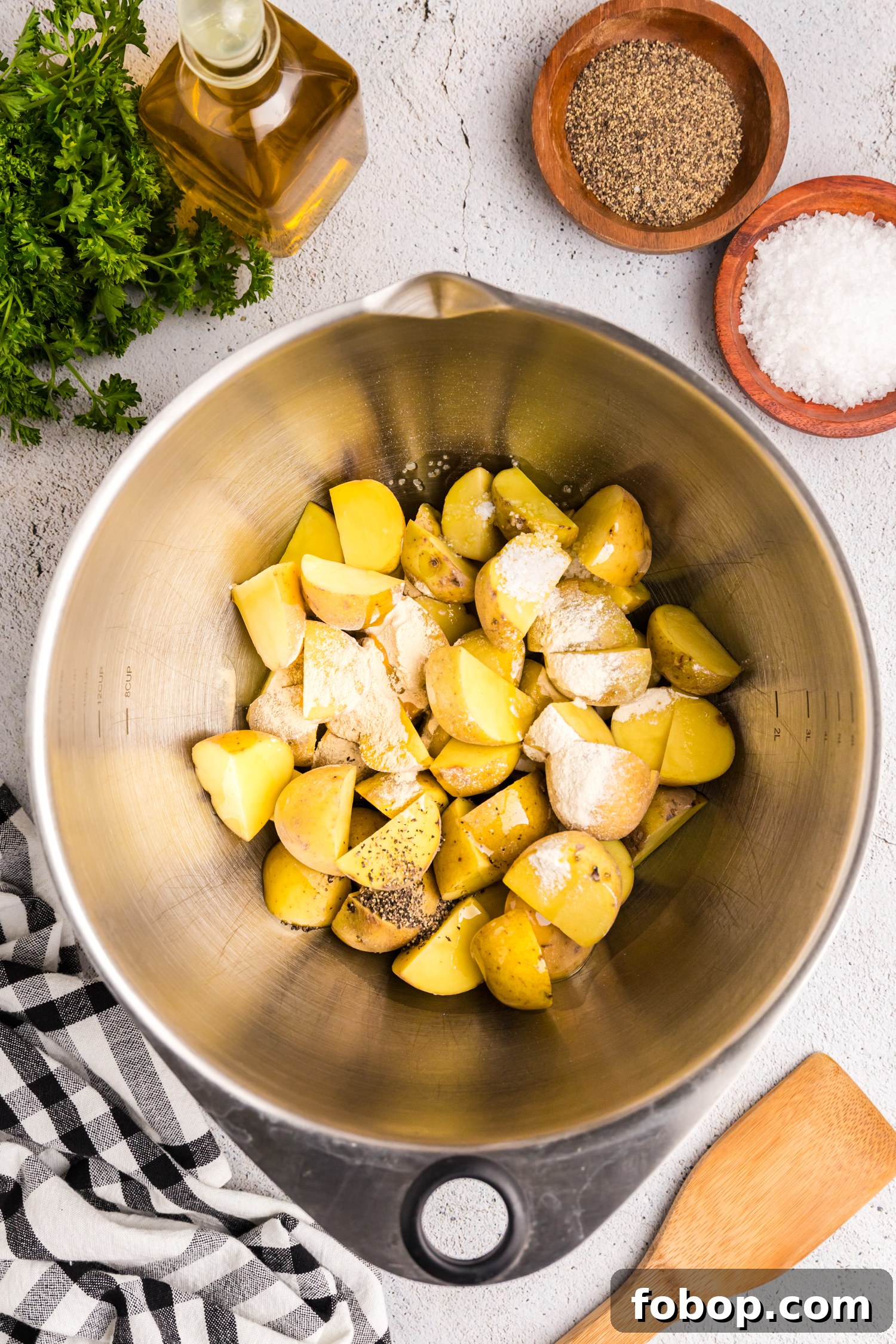 The image shows a stainless steel mixing bowl filled with quartered yellow potatoes seasoned with salt, pepper, and flour. The bowl is placed on a light-colored textured surface, surrounded by ingredients including a bottle of olive oil, wooden bowls containing salt and black pepper, and a bunch of fresh parsley. A wooden spatula and a black-and-white checkered kitchen towel are also present in the scene, suggesting a preparation process for a roasted or fried potato dish.