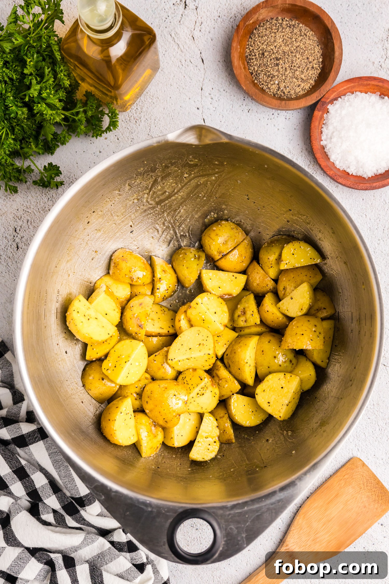 A stainless steel mixing bowl filled with quartered yellow potatoes that have been evenly coated with oil, salt, and black pepper. The bowl is placed on a light-colored textured surface, with ingredients such as a bottle of olive oil, wooden bowls containing salt and black pepper, and a bunch of fresh parsley surrounding it. A wooden spatula and a black-and-white checkered kitchen towel are also in the scene, indicating preparation for a roasted or fried potato dish.