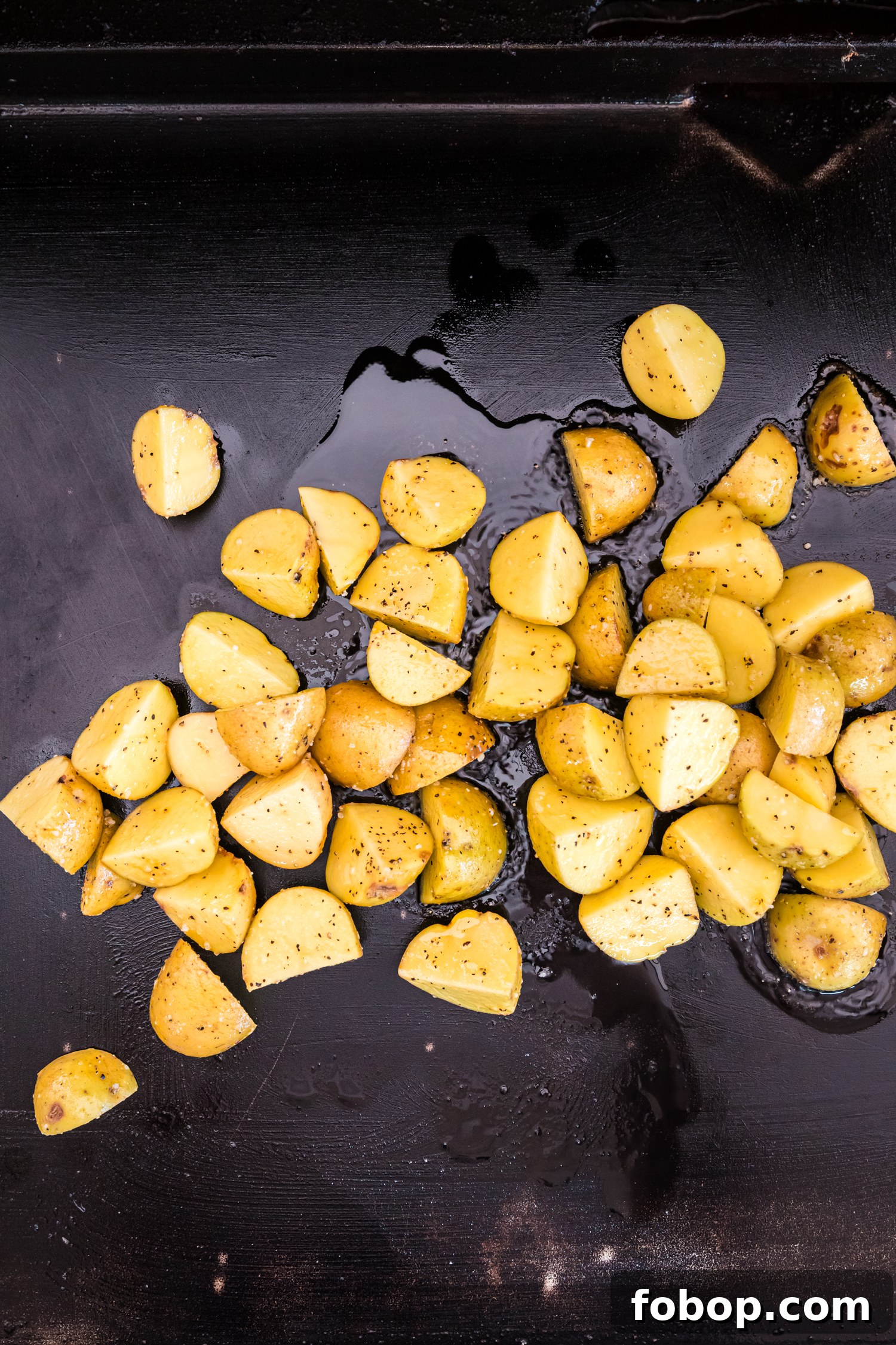 Golden yellow potato pieces, seasoned with salt and black pepper, are spread out on a hot black griddle with a thin layer of oil. The potatoes are beginning to cook, with some edges slightly crisping. The griddle surface has a glossy sheen from the oil, reflecting light. The image captures the early stages of frying the potatoes, likely leading to a crispy, golden-brown texture.