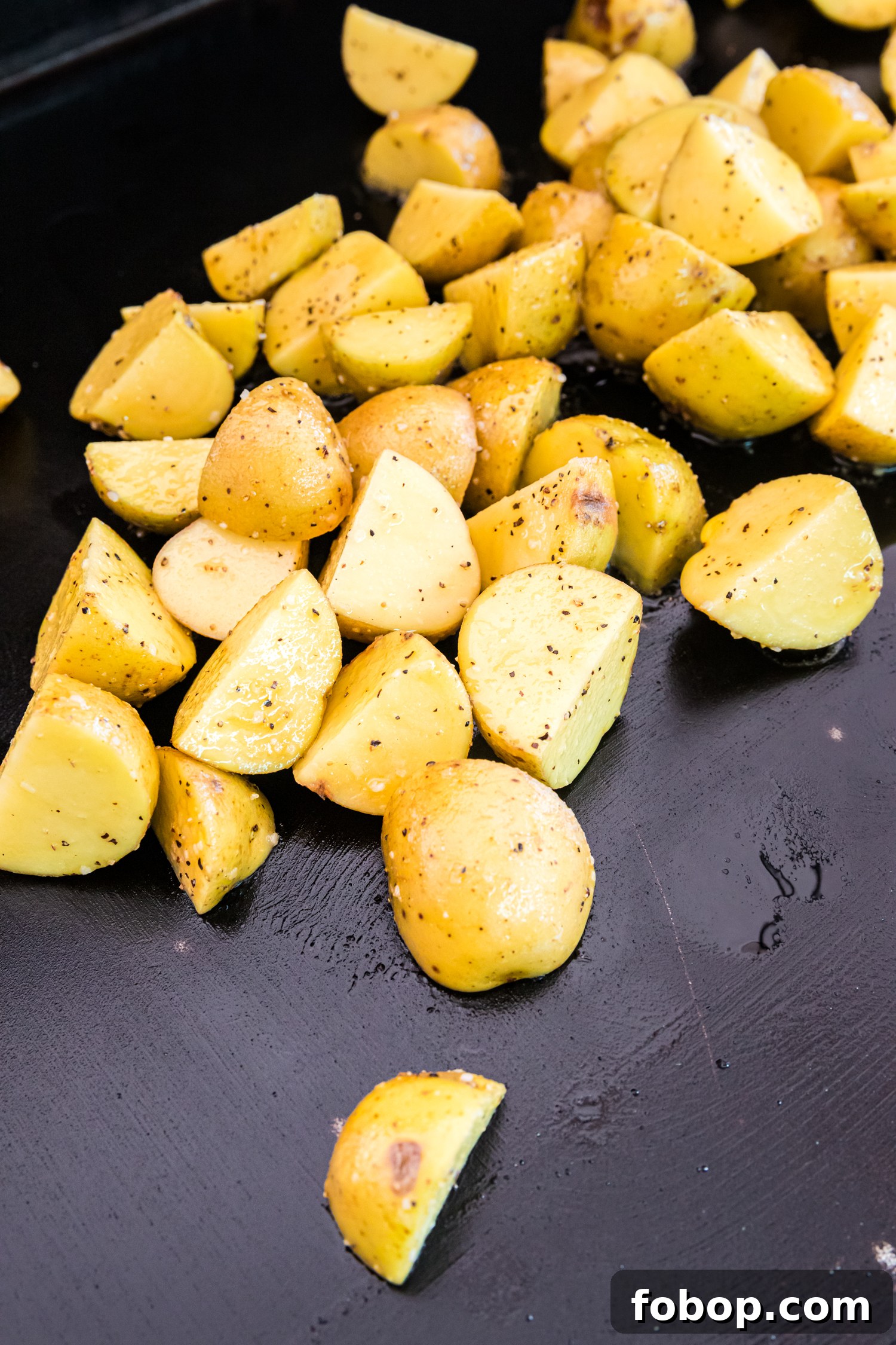 Golden yellow potato wedges are sizzling on a black griddle, coated in oil, salt, and black pepper. Some pieces show the beginnings of a crispy, golden-brown crust. The surface of the griddle has a slight sheen from the oil, enhancing the roasting process. The potatoes are evenly spread out, allowing them to cook thoroughly and develop a delicious crispy texture.