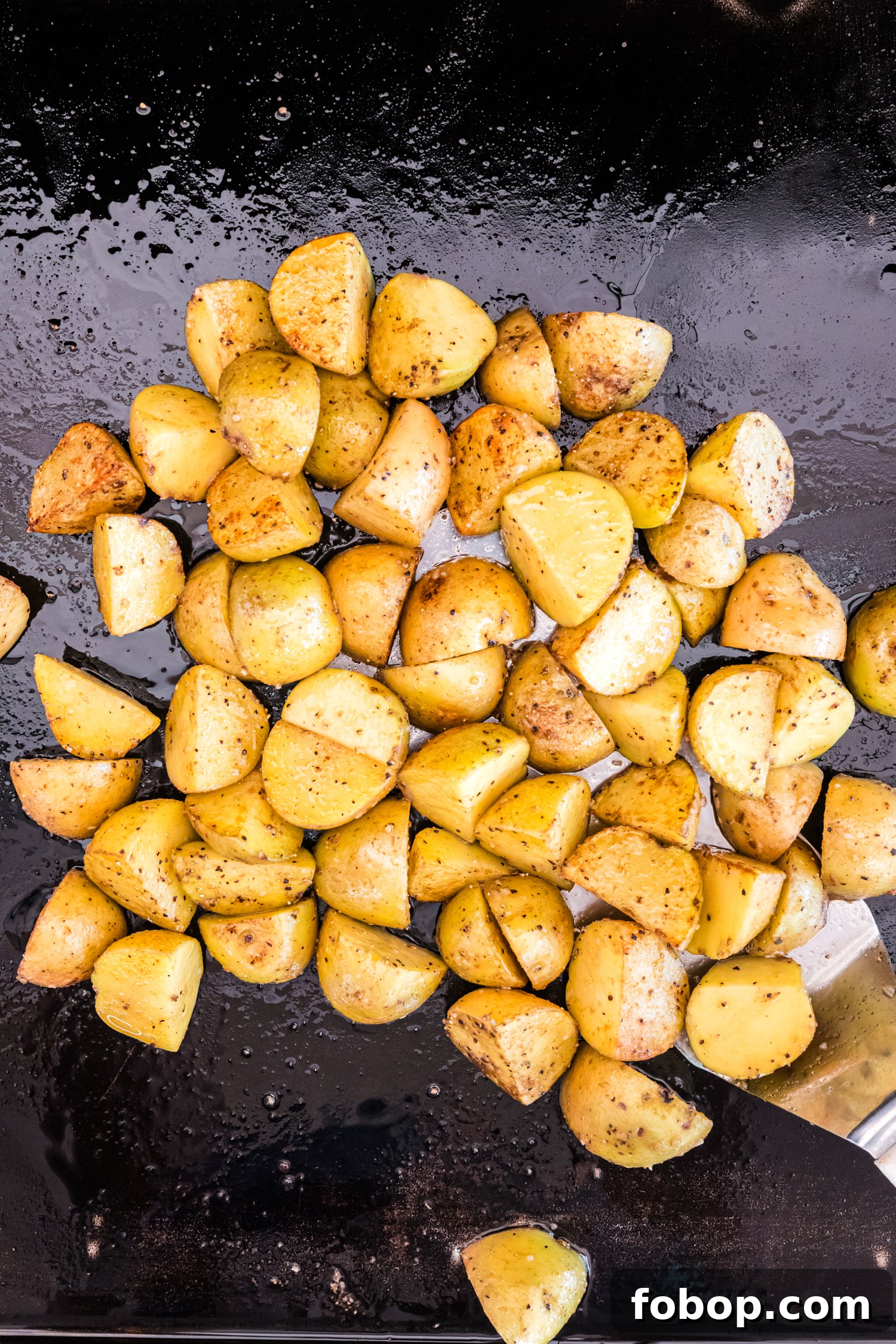 A batch of golden yellow potato wedges is sizzling on a well-oiled black griddle. The potatoes are seasoned with salt, pepper, and other spices, developing a crispy, golden-brown exterior. A metal spatula is partially visible on the right side, ready to flip or stir the potatoes for even cooking. Small droplets of oil and moisture scatter across the griddle surface, enhancing the glossy sheen of the potatoes as they continue to cook.