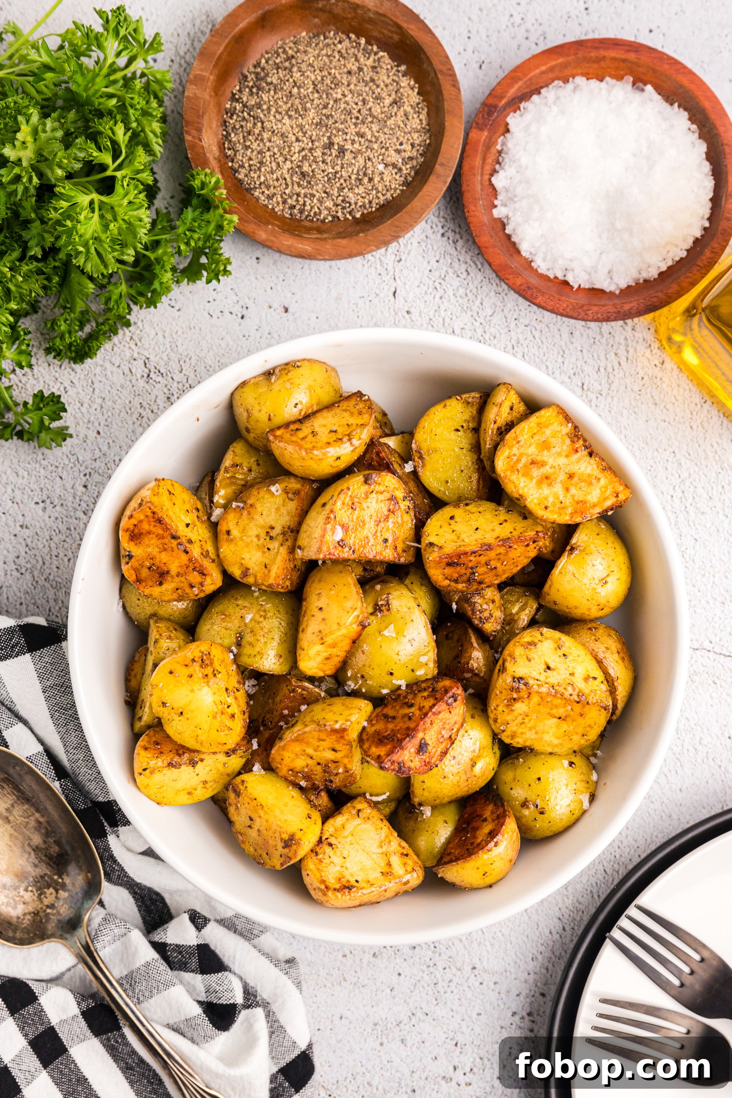 A white bowl filled with golden-brown, crispy fried potatoes sits on a textured surface. The potatoes are seasoned with salt and black pepper, with a slightly charred and crispy exterior. Around the bowl, wooden dishes hold coarse salt and cracked black pepper, a bottle of olive oil is partially visible, and a bunch of fresh parsley adds a touch of green. A black-and-white checkered napkin, a vintage spoon, and stacked plates with forks complete the inviting presentation.