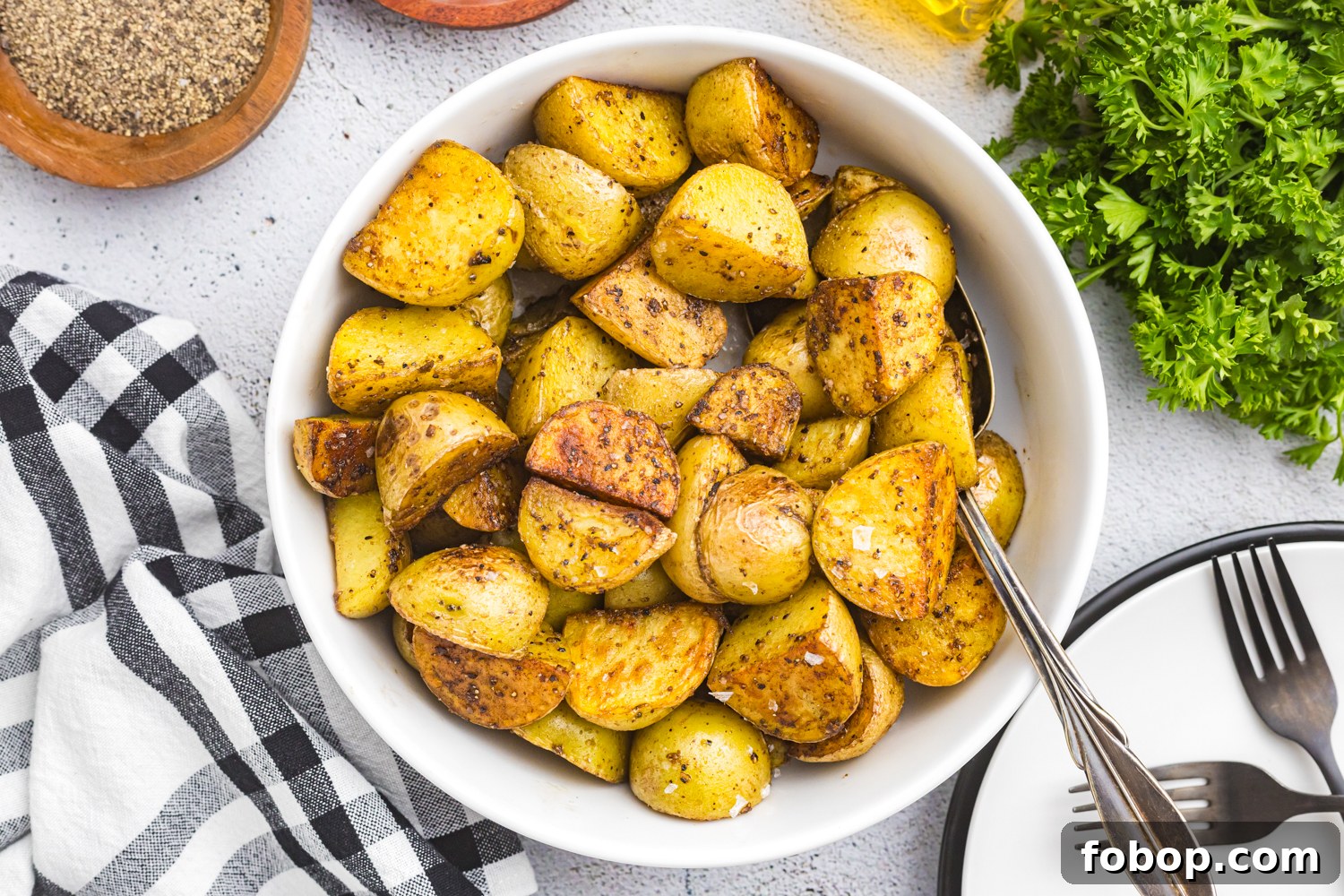 A white bowl filled with crispy, golden-brown fried potatoes sits on a textured surface. The potatoes are well-seasoned with salt, black pepper, and spices, showcasing a crispy exterior and tender interior. A vintage spoon rests in the bowl, ready for serving. Surrounding the bowl, wooden dishes contain coarse salt and cracked black pepper, while fresh parsley adds a touch of green. A black-and-white checkered napkin, stacked plates, and forks complete the inviting and rustic presentation.