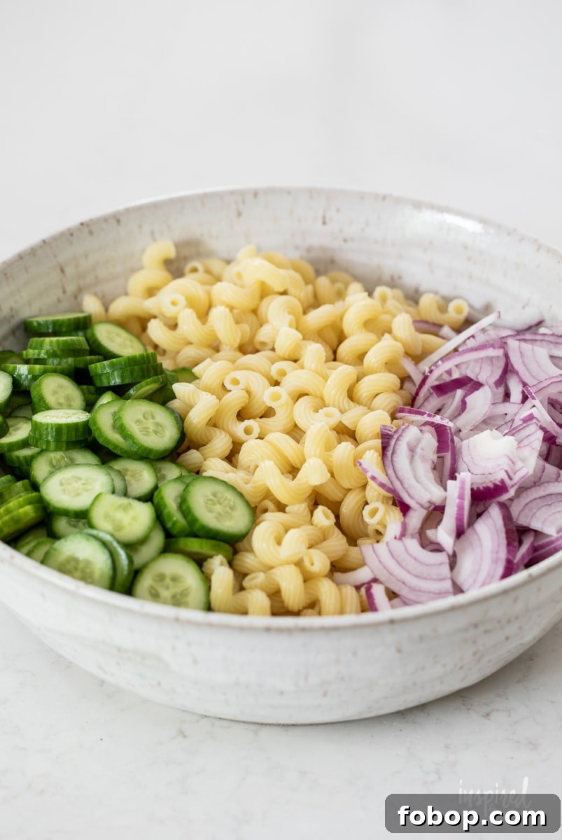 a large bowl with sliced cucumbers, cooked pasta, and red onions.