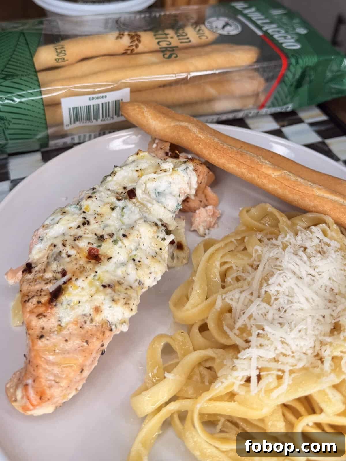 Lemon Ricotta-Stuffed Salmon with Herbed Citrus Sauce 2 A plate with baked salmon topped with creamy cheese and herbs, fettuccine pasta sprinkled with grated cheese, and a breadstick. In the background, a package of Italian breadsticks is visible.