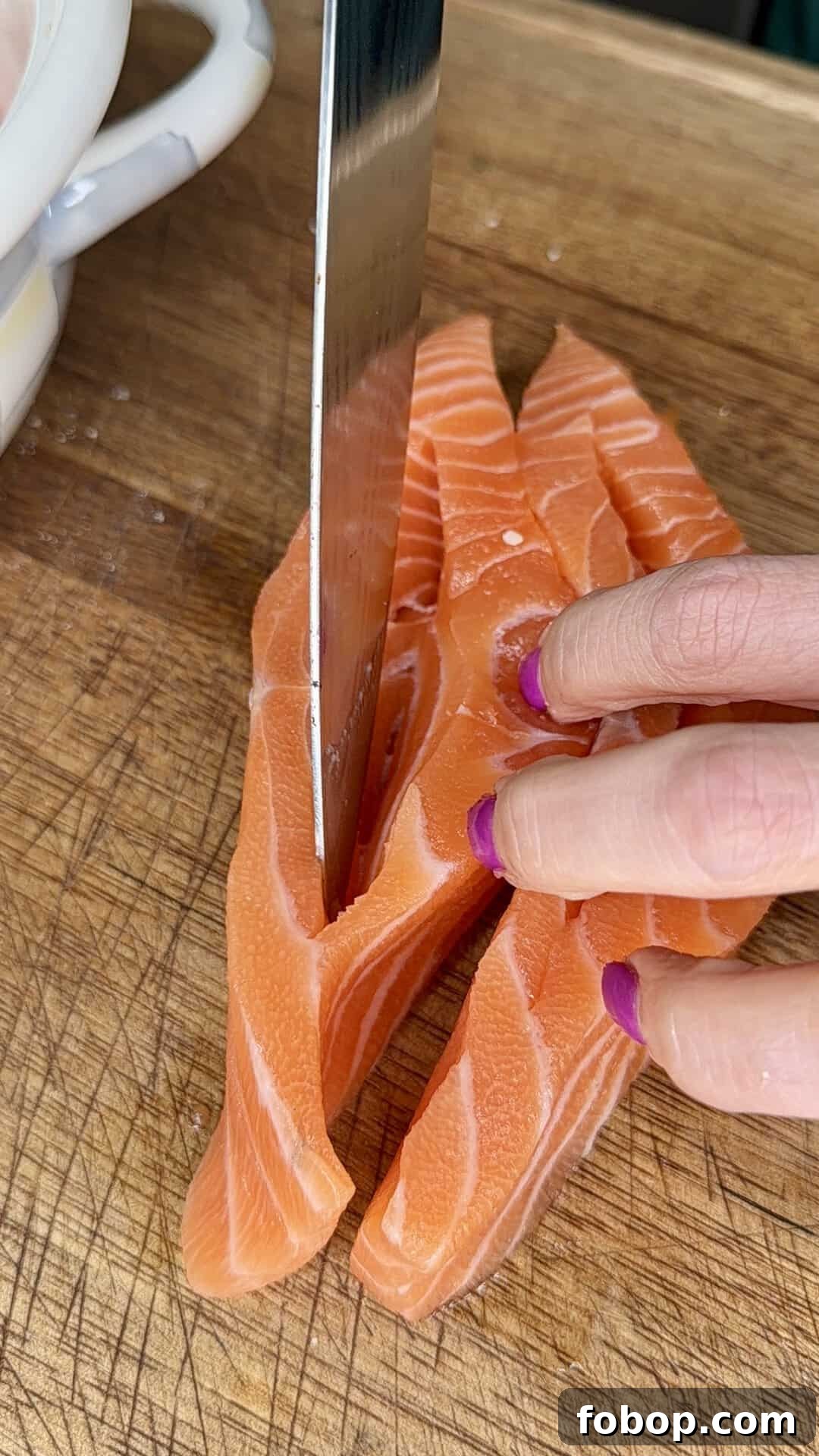 Lemon Ricotta-Stuffed Salmon with Herbed Citrus Sauce 4 A hand with purple-painted nails holds raw salmon while a large knife slices it into thin strips on a wooden cutting board.