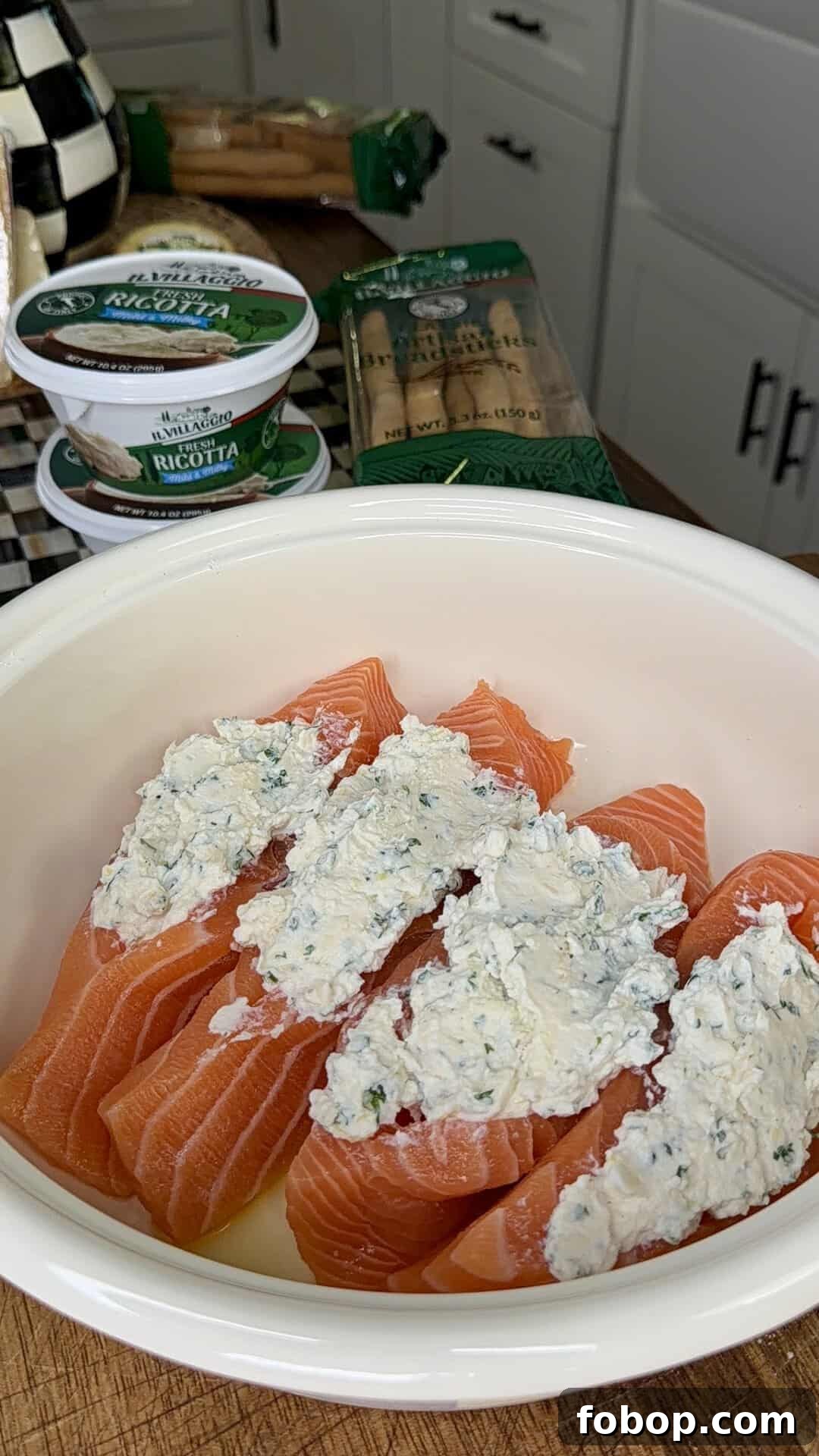 Lemon Ricotta-Stuffed Salmon with Herbed Citrus Sauce 5 A white bowl with raw salmon fillets split open and stuffed with a creamy ricotta and herb filling. Containers of ricotta cheese and packages of cannelloni pasta are on the counter in the background.