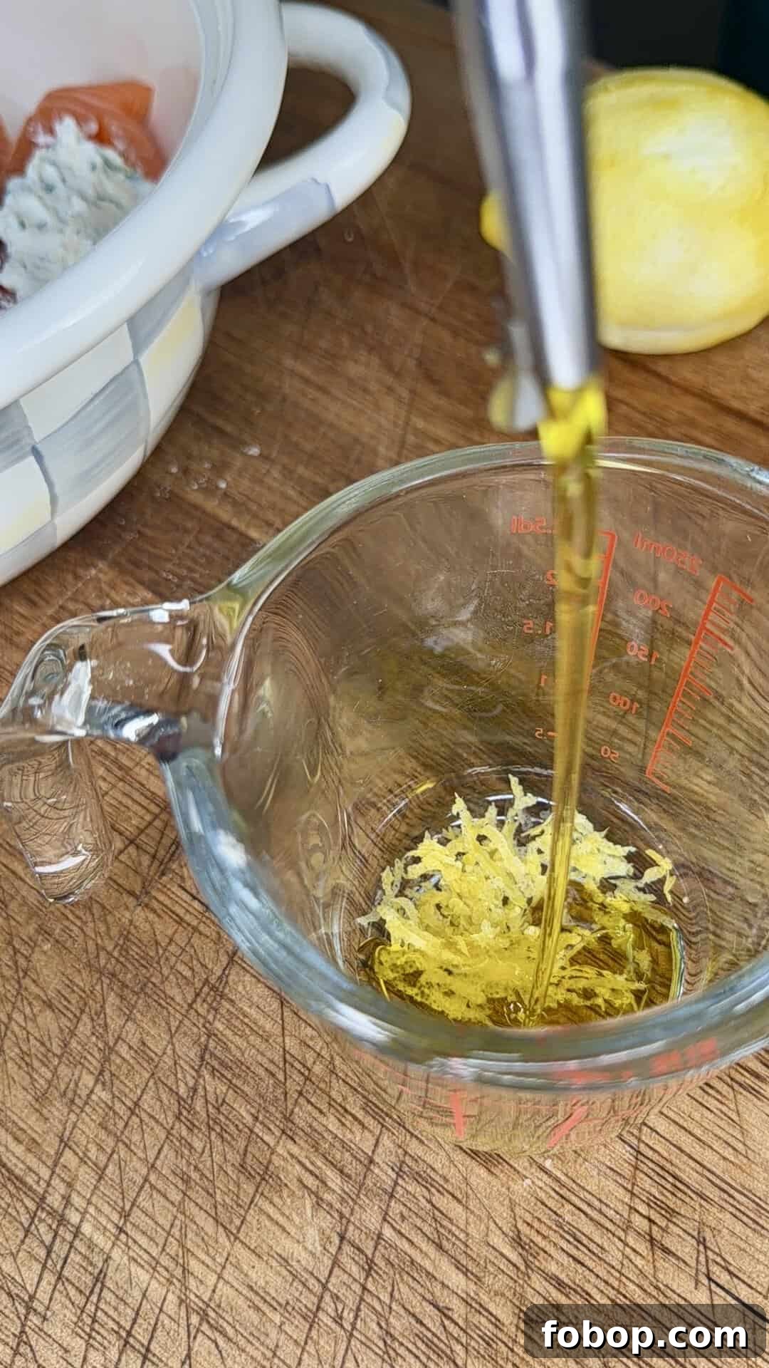Lemon Ricotta-Stuffed Salmon with Herbed Citrus Sauce 6 Olive oil being poured into a glass measuring cup containing lemon zest, on a wooden cutting board. A lemon and a bowl with cheese and tomato are visible in the background.