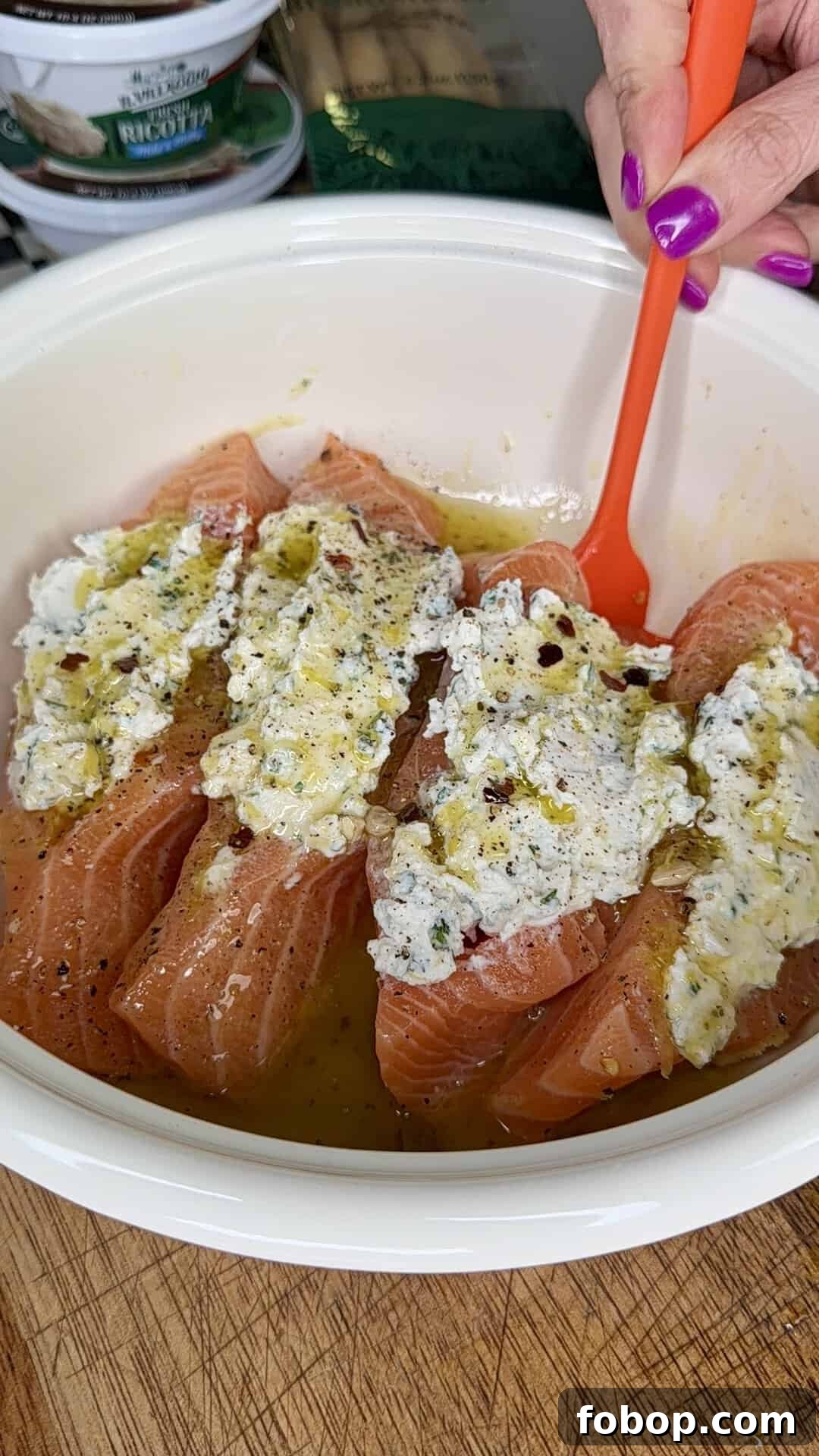 Lemon Ricotta-Stuffed Salmon with Herbed Citrus Sauce 7 Four raw salmon fillets in a white bowl are topped with a creamy herb mixture and oil. A hand with purple nail polish holds an orange spoon next to the salmon.
