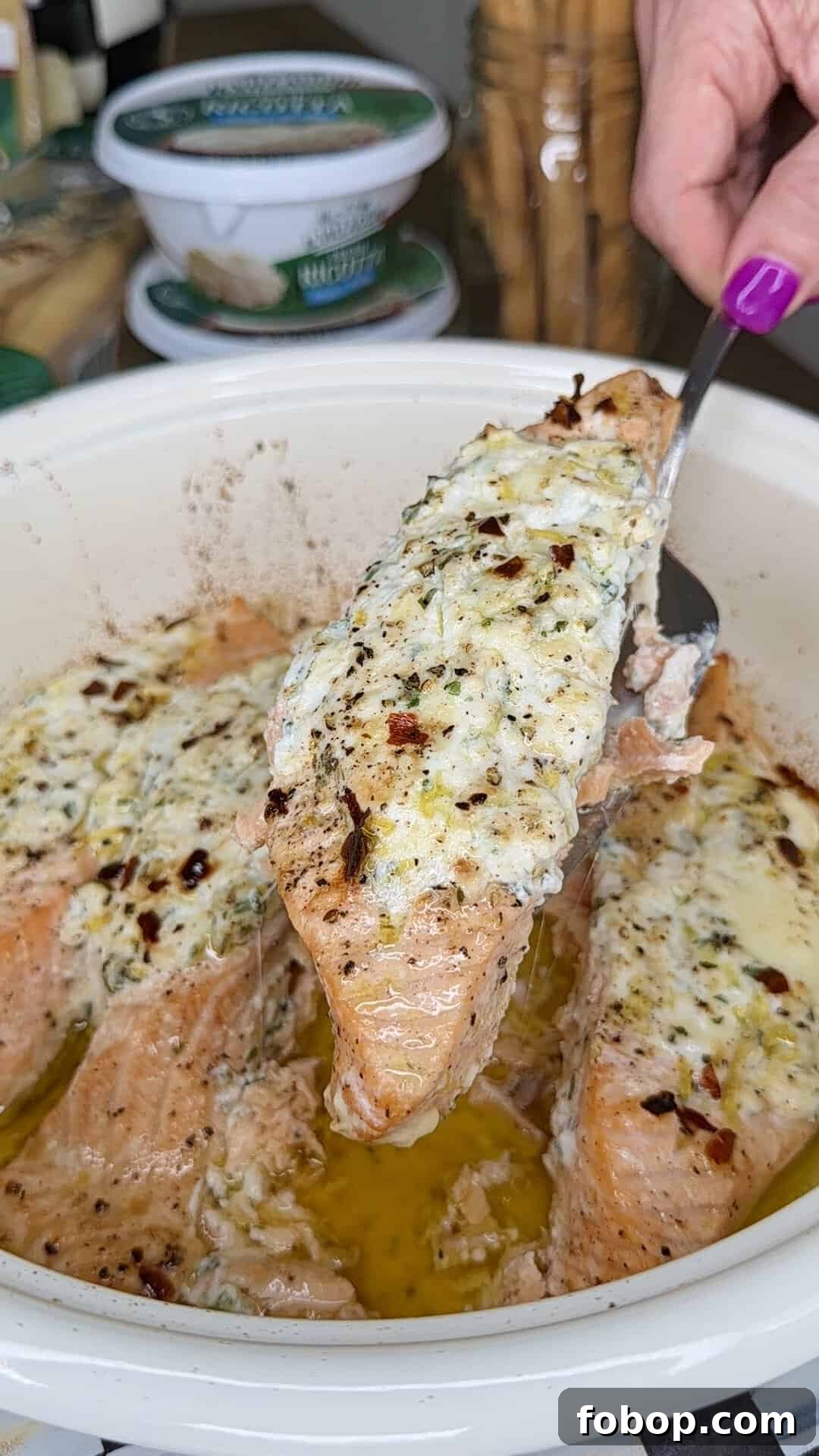 Lemon Ricotta-Stuffed Salmon with Herbed Citrus Sauce 9 A hand with purple nail polish lifts a piece of baked salmon topped with creamy herb and cheese mixture from a dish, with more salmon pieces and melted butter visible below.