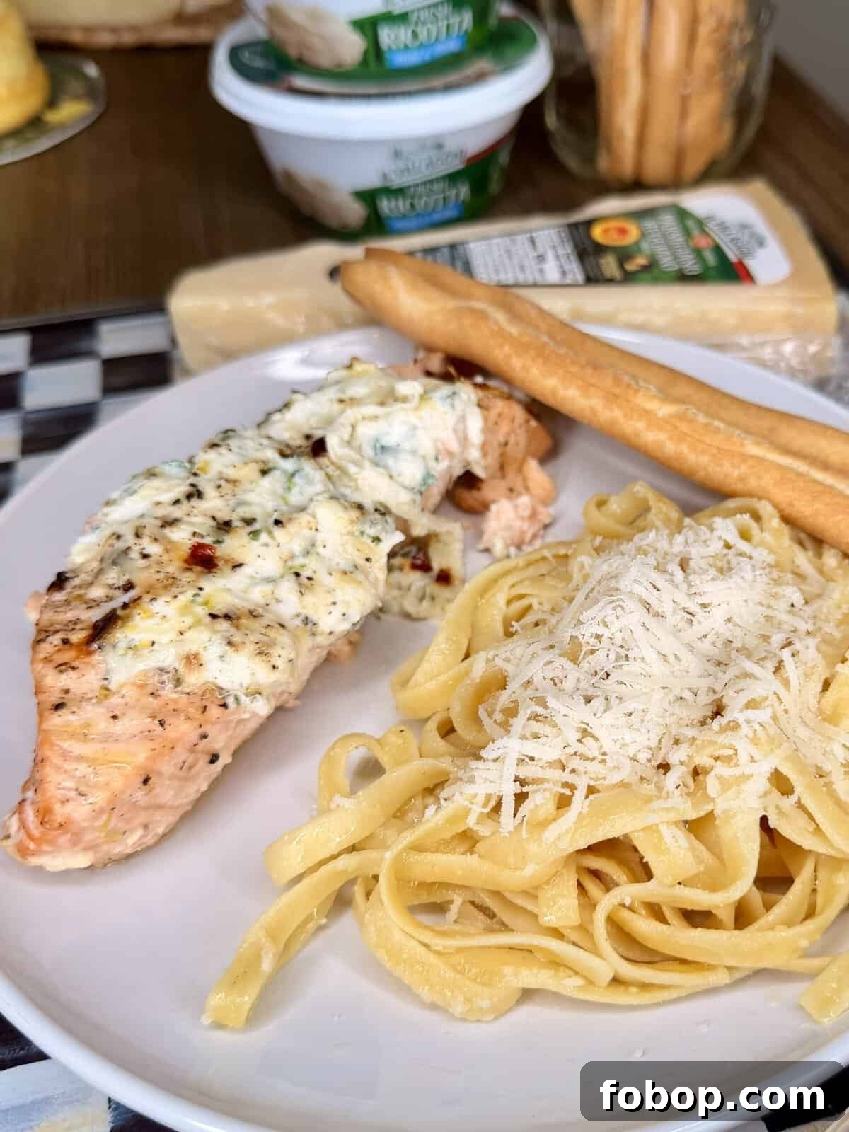 Lemon Ricotta-Stuffed Salmon with Herbed Citrus Sauce 10 A plate with creamy baked salmon, a breadstick, and fettuccine pasta topped with grated cheese. In the background are cheese packages, ricotta containers, and more breadsticks on a table.