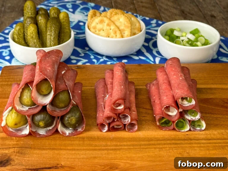Savory Dried Beef Roll-Ups Recipe for Appetizers and Snacks 2 Stacks of variations of dried beef roll-ups on a wooden cutting board with a bright blue napkin and small white dishes of dill pickles, crackers and green onion slices in the background.